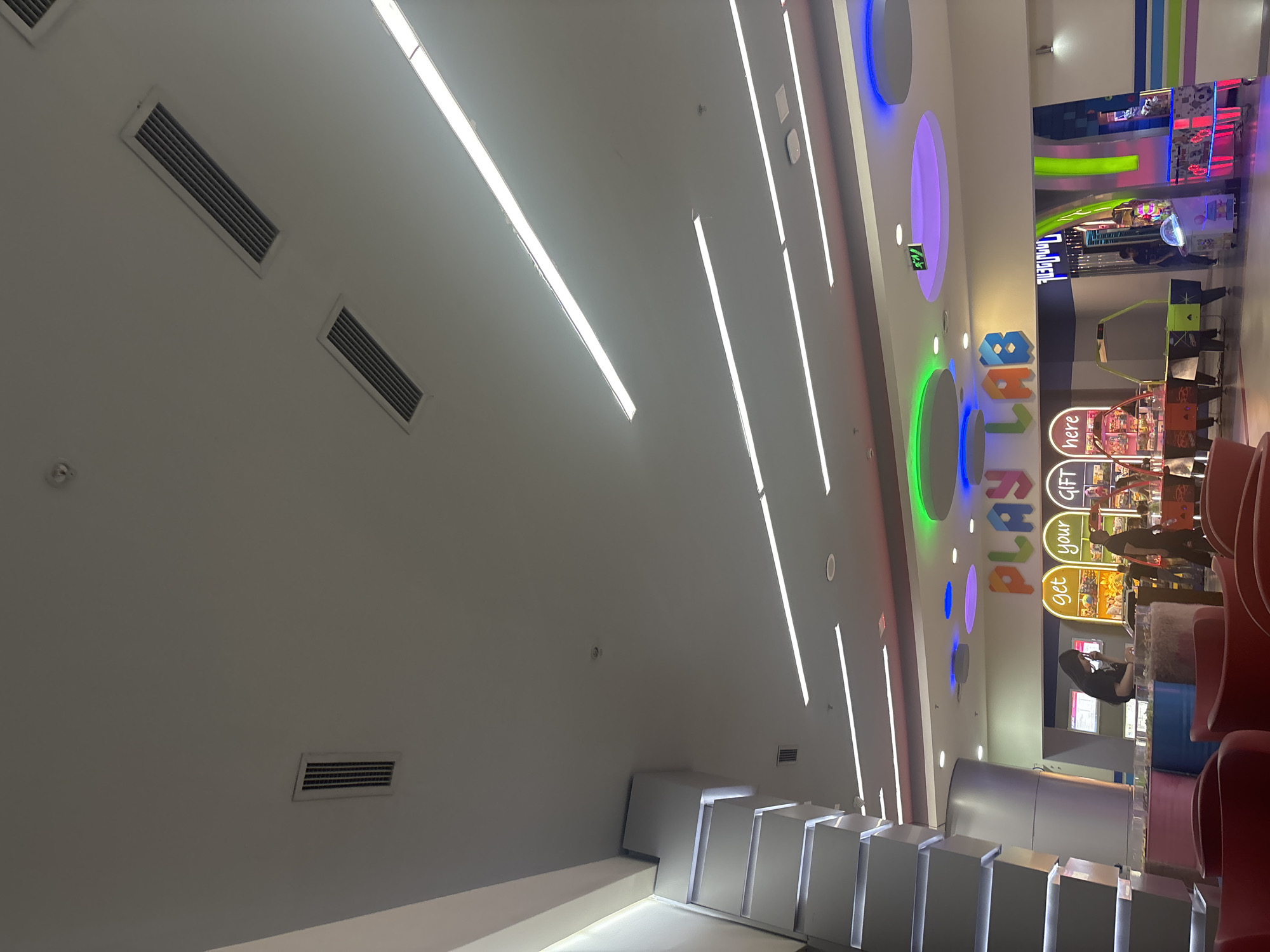 The image captures an indoor entertainment or gaming area, likely within a mall, with bright artificial lighting and colorful decor. The perspective is tilted, looking towards a wall featuring a prominent "PLAY LAB" sign with multicolored letters.
The ceiling, which takes up a significant portion of the left side of the frame, is white and features multiple long, linear fluorescent or LED light fixtures running parallel to each other. Several rectangular air vents are also visible on the ceiling.
On the right side of the image, where the "PLAY LAB" sign is located, the wall is adorned with large, circular light installations in shades of blue, purple, green, orange, and yellow, creating an vibrant atmosphere. Below the "PLAY LAB" sign, several arcade and prize machines are visible. Two stylized, bright green racing car-like game units are prominent in the mid-ground.
Several individuals are present in the scene. One person with long dark hair, wearing a black top, is seated at a counter on the far right, facing away from the camera, seemingly engaged with a device or item on the counter. Other people are partially visible further back, near the gaming machines and a "get your gift here" counter, suggesting interaction with the games or prize redemption.
In the foreground on the left, there's a modern, tiered seating or display structure made of white, angular blocks. The overall impression is that of a modern, lively arcade or family entertainment center. Visible text includes "PLAY LAB" in large letters, "get your gift here" on a prize counter, and partially visible text like "DOUBL..." and "check out" on arcade machines. The setting is clearly indoors, illuminated by artificial light.
