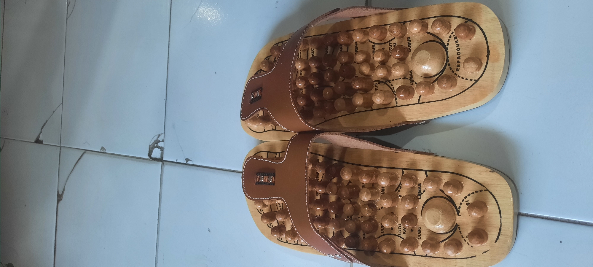 A pair of brown leather and wood acupressure sandals are displayed on a light blue tiled floor. The sandals are oriented vertically, with the soles facing upwards and the toe straps pointing towards the top of the image. Each sandal sole is made of wood and features a grid of small, rounded wooden pegs, intended for acupressure. Black text, written in Indonesian, is printed on the wooden soles, indicating various acupressure points. A smooth, rounded wooden protrusion is present near the heel of each sandal. The background consists of the tiled floor, with visible grout lines and some minor imperfections or damage to the tiles. The lighting appears to be from above, casting subtle shadows. The scene is static and appears to be an indoor setting.