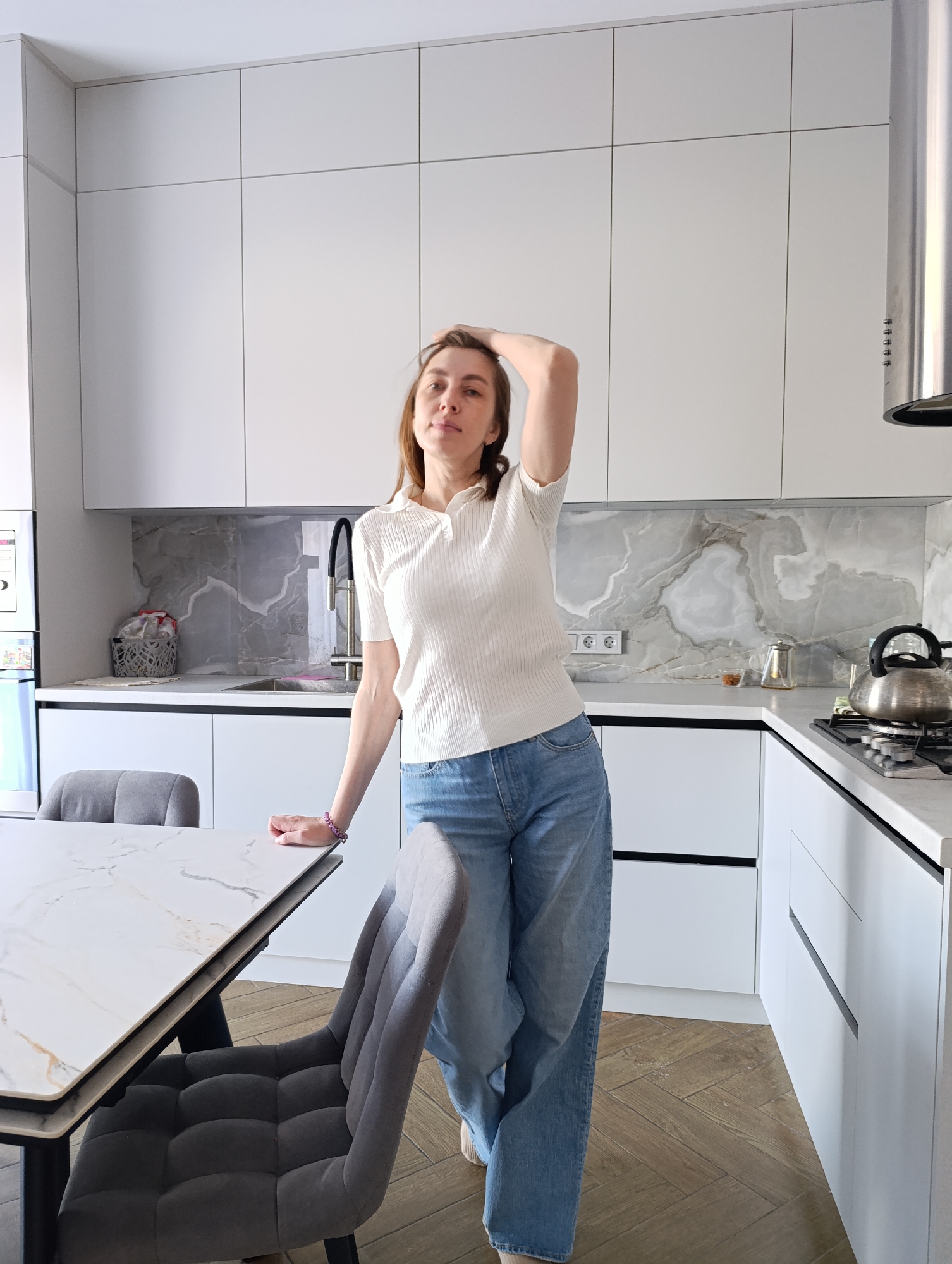 A young adult woman is captured in a full-length portrait within a modern kitchen and dining area in Chaikovsky, Russia. She stands centrally, facing the camera with a neutral expression, her left hand gently placed on her head and her right arm resting on a white marble-patterned dining table. She is dressed in a short-sleeved white ribbed polo shirt and loose-fitting blue jeans.
The kitchen features a clean, minimalist design with white flat-panel cabinetry. Upper cabinets line the wall, while lower cabinets have sleek black integrated handles. A light grey marble-patterned backsplash adds visual interest above the light grey countertops. To the left, a black flexible faucet is visible over a sink area, with a grey woven basket and a white electrical outlet nearby. On the right side of the kitchen, a silver kettle sits on a gas stovetop, next to a small glass teapot and a dish containing nuts or seeds. A cylindrical stainless steel range hood is prominently positioned above the cooking area.
Around the dining table, two grey upholstered chairs with tufted backs are partially visible. The floor throughout the space is covered with light brown wooden planks arranged in a herringbone pattern. The scene is brightly lit, indicating it is daytime. No visible text is present in the image.
