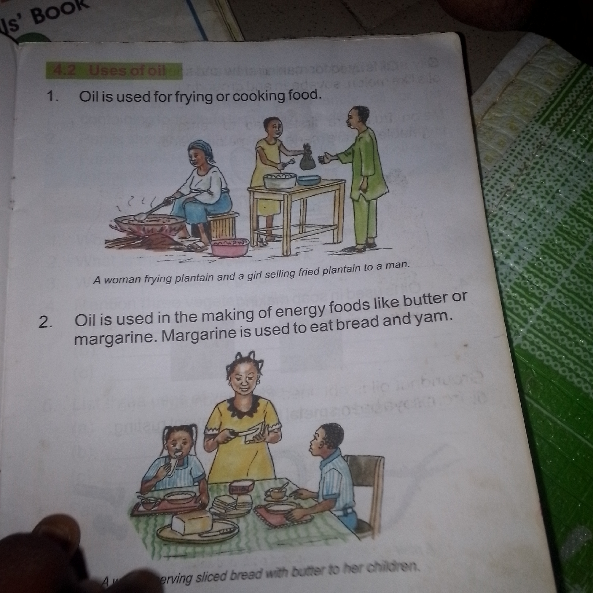 The image is a page from a book, likely a textbook, as indicated by the numbered sections and illustrations. The page features two illustrations depicting the uses of oil. The top illustration shows three people: a woman frying plantain in a large pot, a girl selling fried plantain to a man, and another woman sitting down. The text below this illustration reads "A woman frying plantain and a girl selling fried plantain to a man."
The second illustration depicts a family at a table. A woman is serving sliced bread with butter to two children, who are eating at the table. The text below this illustration reads, "Oil is used in the making of energy foods like butter or margarine. Margarine is used to eat bread and yam." The overall environment appears to be domestic or a marketplace setting. The image is captured in what appears to be an indoor environment, with a green patterned fabric visible on the right side of the image. The image does not provide any information about a specific city or location.