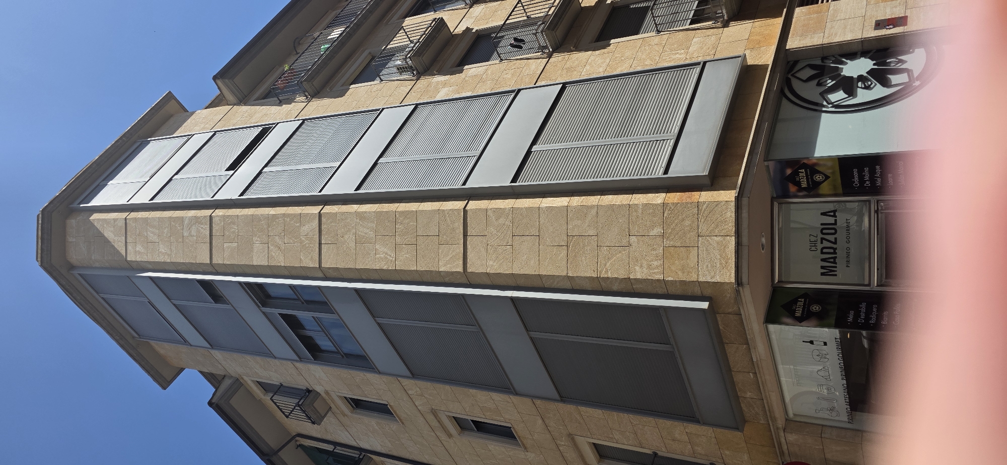 The image shows a multi-story building with a corner facade made of light brown stone blocks and grey metallic paneling. The building has multiple windows with closed grey roller shutters. On the ground floor, there are shopfronts with large glass windows. One of the shopfronts displays signage that reads "CHEZ MADZOLA PIRINEO GOURMET" and lists various products. The street or sidewalk is visible in the foreground, partially obscured by a blurry pink object, possibly a finger. The sky is clear and blue. The architectural style suggests an urban setting, likely in a European city. Based on the signage, this building is located in Huesca, Spain, at the "Chez Madzola Pirineo Gourmet" shop.