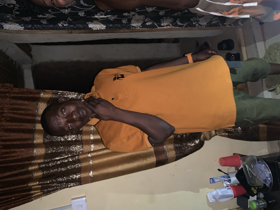 An individual wearing an orange polo shirt rests on a bed with a gold patterned blanket. Their hand is touching their neck. A person lies on the top bunk of what appears to be a bunk bed. There are a few cups and toiletries on a table, and a white electrical socket is visible on the wall. A pair of shoes and a wallet sit on the floor behind the individual in the orange shirt.