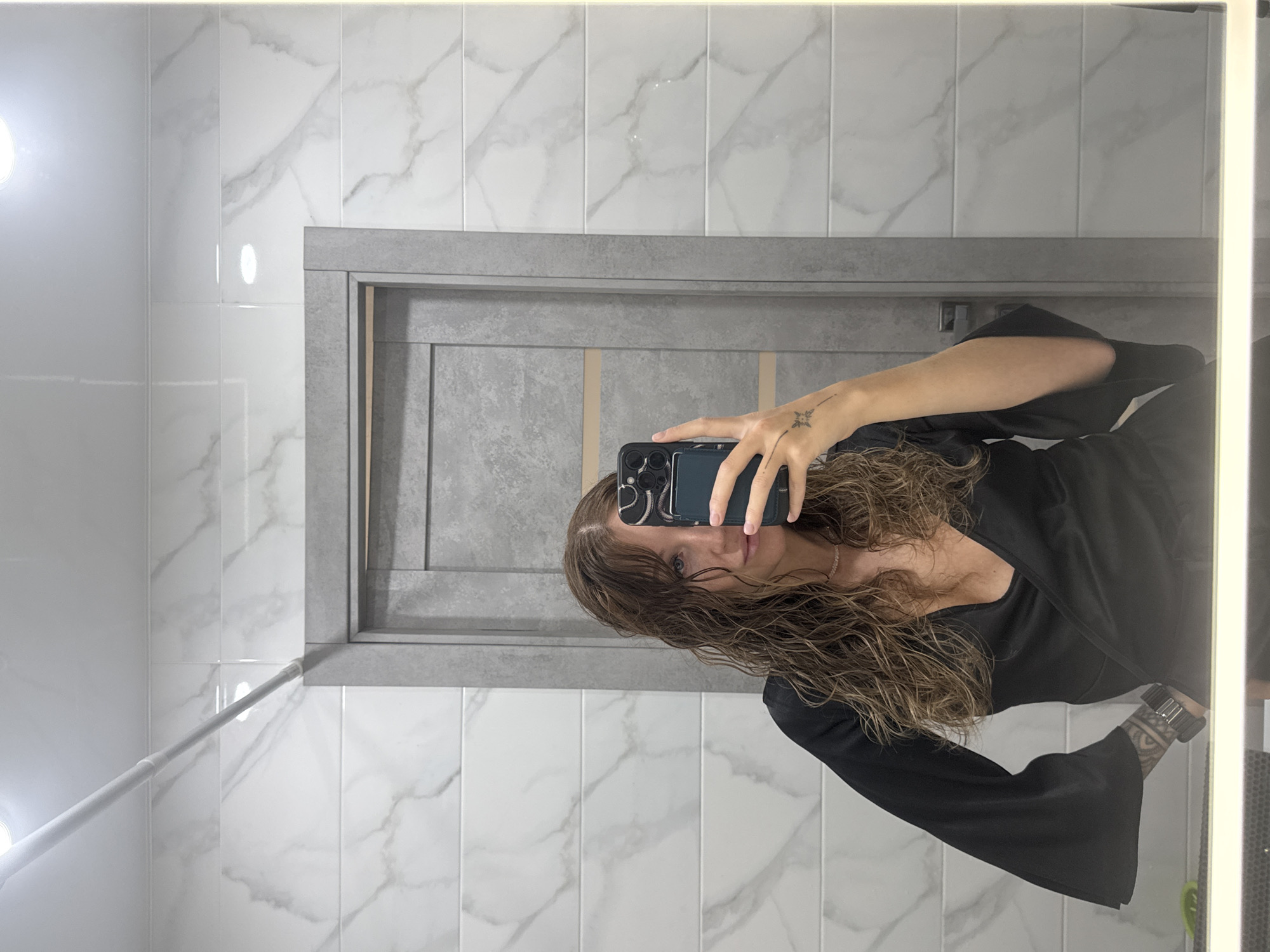 A woman is taking a selfie in a bathroom. The bathroom has white marble-patterned tile walls and a gray door frame. The woman is wearing a black long-sleeved top and holding a phone in her left hand. Her hair is curly and wet. There is a tattoo on her left wrist. The location is Chaikovsky, Russia.