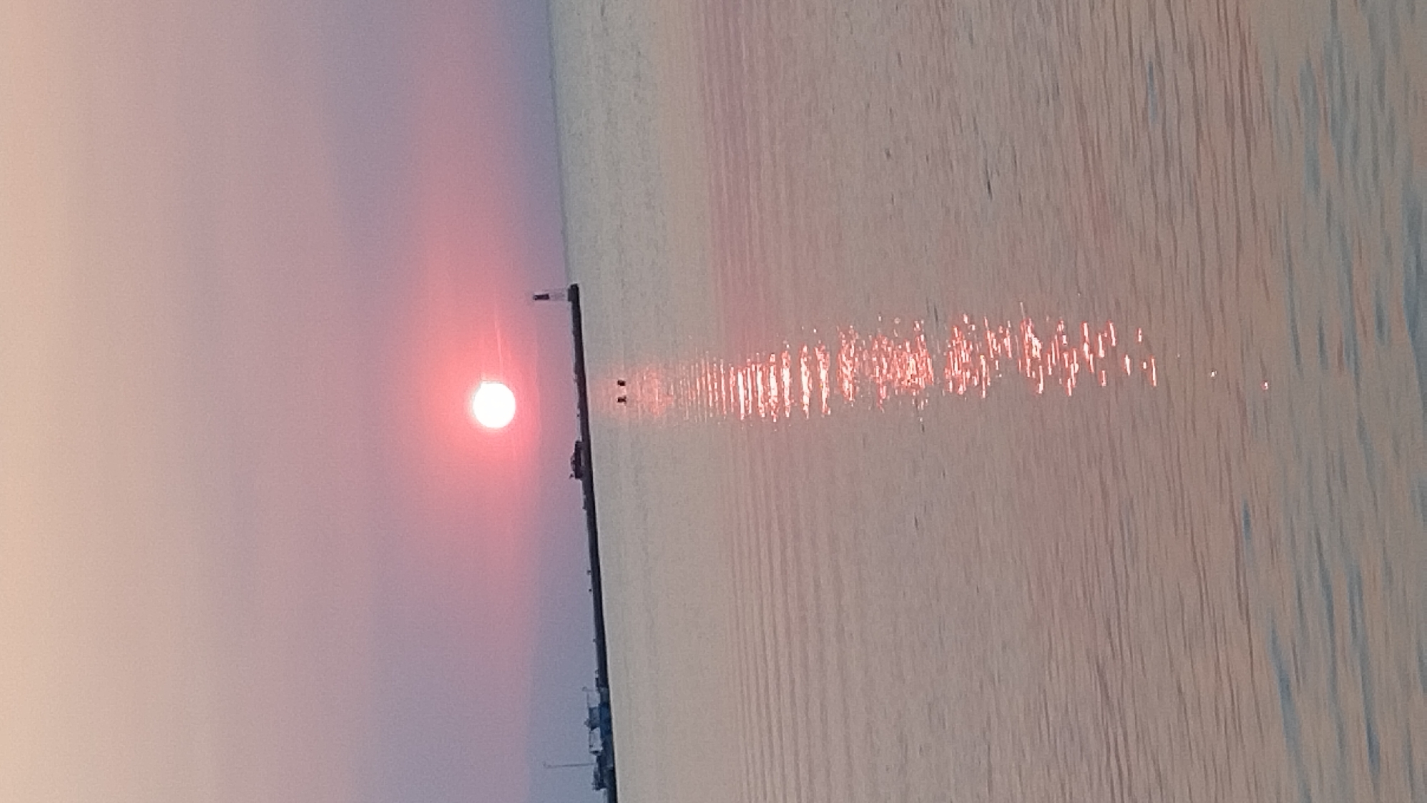 A serene sunset over the Aegean Sea near Neoi Epivates, Greece, is captured in this vertical shot. The sun, a bright, reddish-orange orb, is positioned in the upper-center of the frame, casting a shimmering reflection across the calm water. The sky exhibits a soft gradient of pink and purple hues, transitioning to a lighter tone near the horizon.
In the distance, a long pier or breakwater extends into the sea from the left. Several boats are docked along the pier, and a lighthouse stands at its end. Two figures, appearing as dark silhouettes, are visible in the water near the pier, suggesting they are swimming or wading. The water itself is gently rippled, creating a textured surface that reflects the ambient light. The overall scene conveys a sense of tranquility and natural beauty.