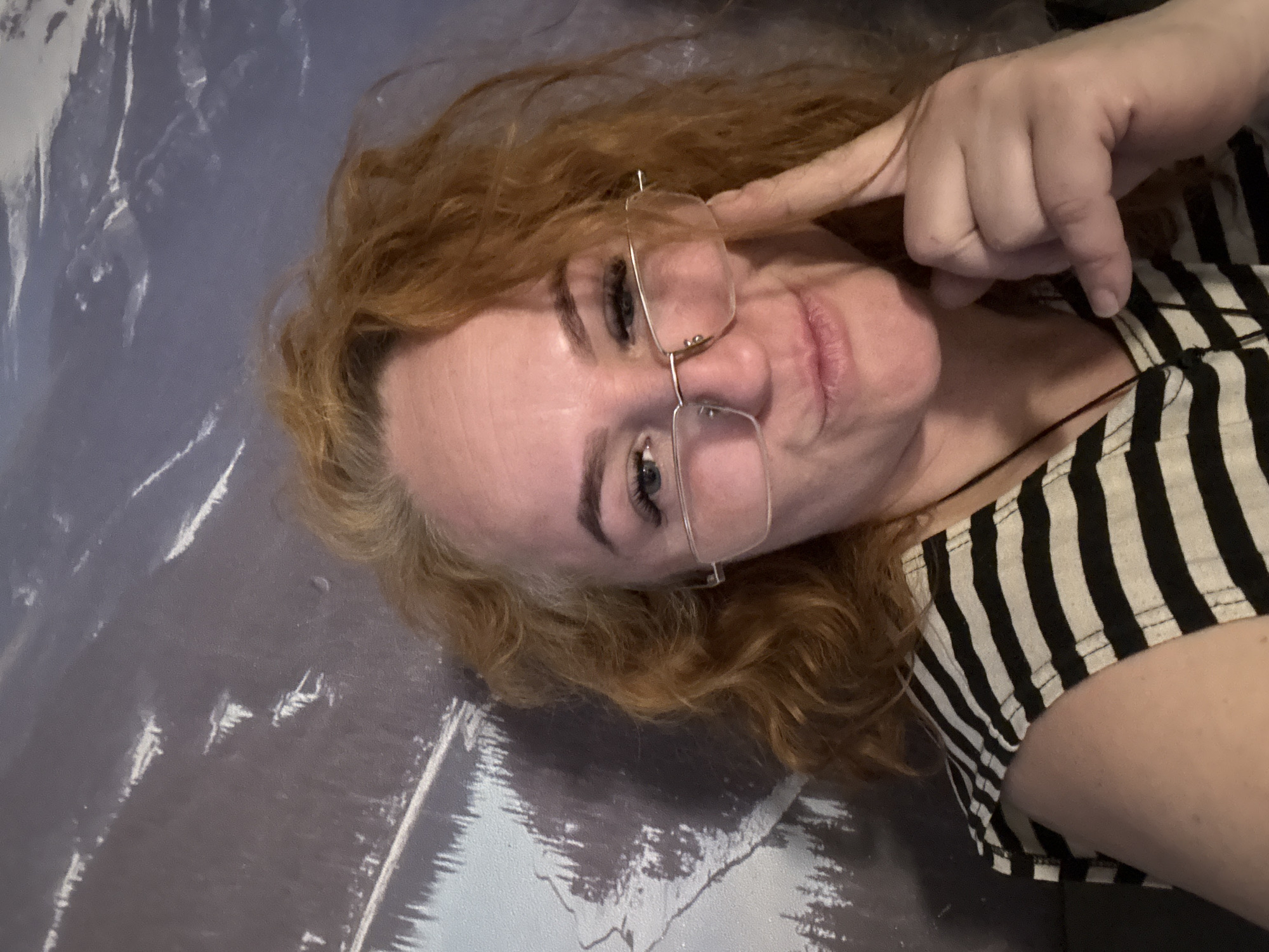 A woman with auburn, curly hair is lying on a surface with a gray and white mottled pattern. She is wearing glasses and a black and white striped shirt. Her right index finger is touching the frame of her glasses. A thin black string is visible around her neck. The location in the city of Chaikovsky, Russia is not discernible from the image.