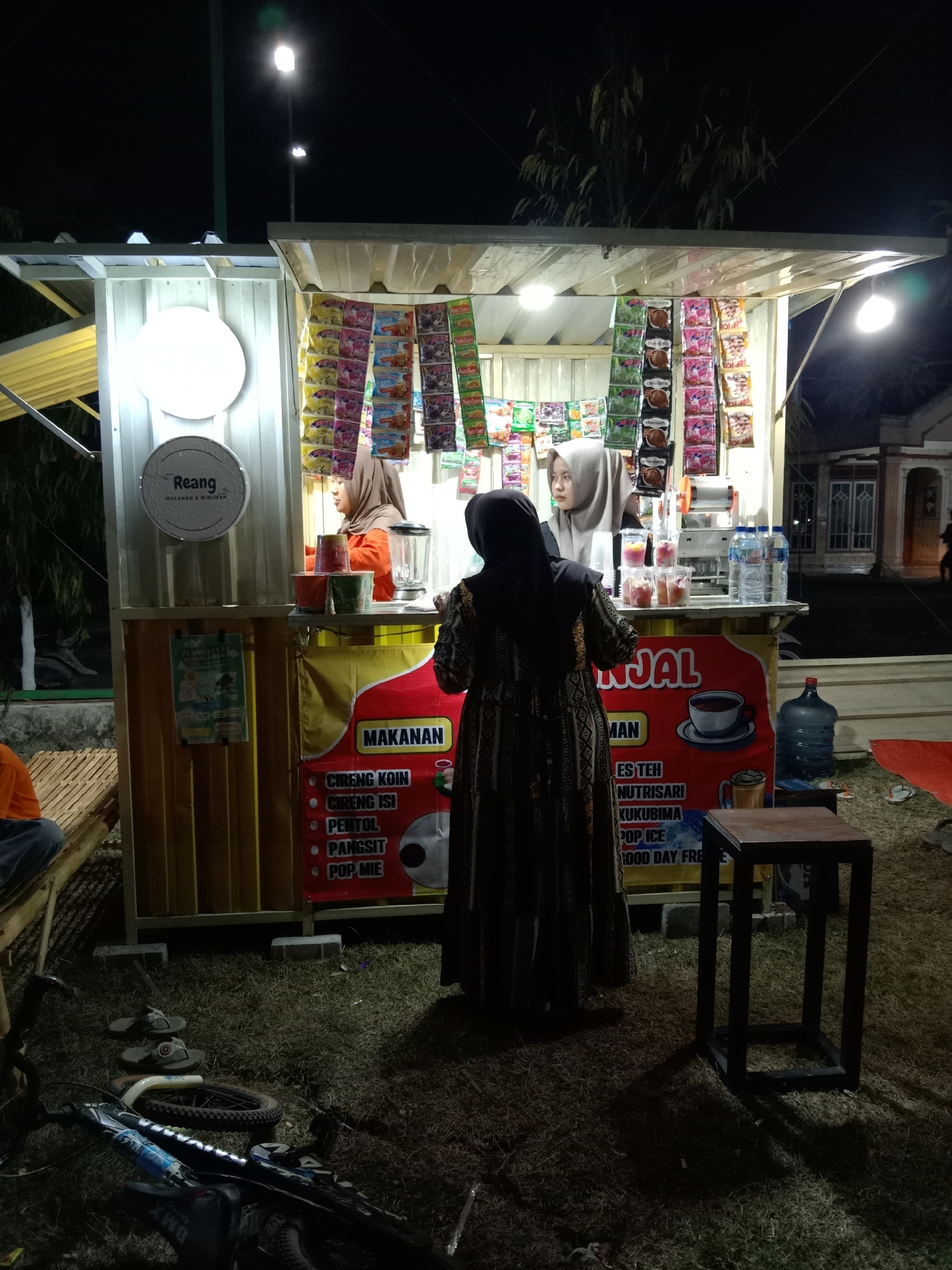 In Stiaji, Indonesia, at night, a food and drink stall is set up. Two women, one in an orange shirt and headscarf and another in a black abaya and headscarf, are working behind the counter. The woman in the orange shirt is making drinks, while the woman in the abaya is facing away from the camera, seemingly interacting with a customer. The stall displays various packaged snacks and drinks. A bicycle and a pair of flip-flops are on the ground in front of the stall. A small wooden stool is positioned to the right of the stall. The surrounding area is dimly lit, with trees and a building visible in the background.