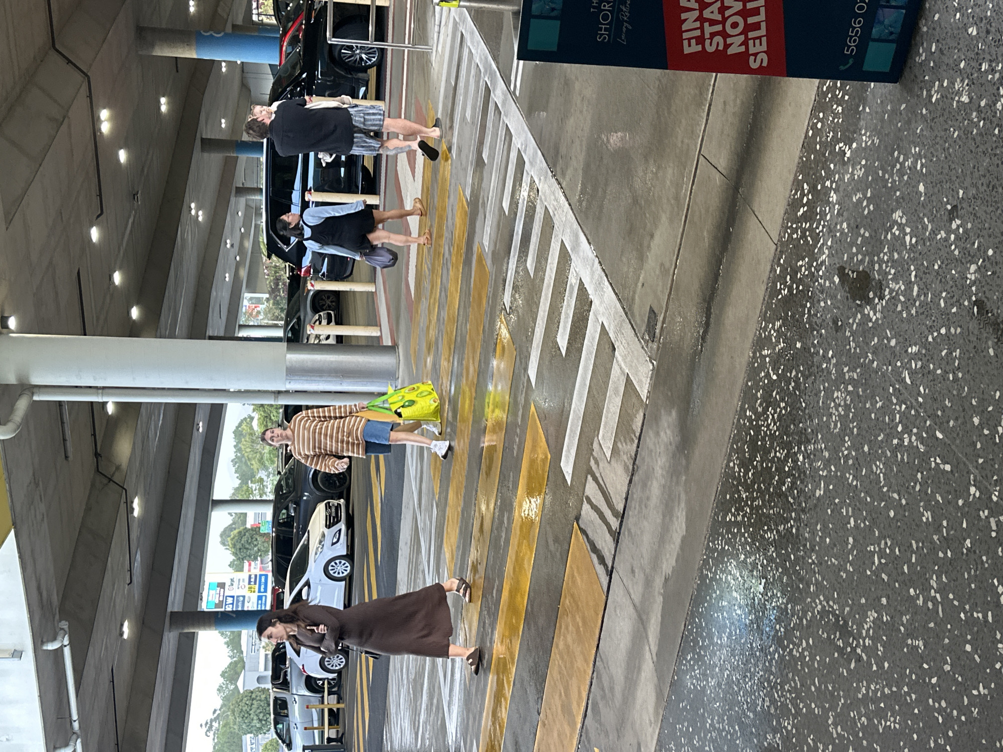 The image depicts a pedestrian crossing in a parking lot, possibly at the Coffs Harbour Jetty, Australia. Several people are visible crossing the zebra stripes, which appear wet and reflective, indicating recent rain.
In the foreground, a woman wearing a long brown dress and sandals walks across the crossing. To her right, a person in a striped long-sleeved shirt and blue shorts walks while carrying a bright yellow tote bag. Further in the background, a man with short hair wearing a black shirt and striped shorts is seen holding a young child. Behind them, a woman in a dark dress and white top is walking.
The environment includes a covered parking structure with overhead lighting. Several cars are parked in the background, including a white hatchback, a black SUV, and a silver ute. A sign with the text "THE SHORE Luxury Retreat" and "FINAL STAGE NOW SELLING" is partially visible on the right side of the image. The ground surface is a mixture of wet concrete and a speckled aggregate material.