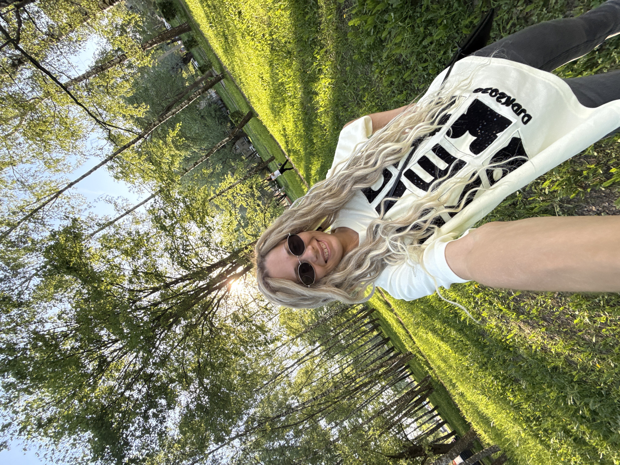 The image captures a selfie taken by a blonde woman in an outdoor setting. She is wearing round sunglasses, a white t-shirt, and black pants, lying on a grassy area. The t-shirt has a black graphic design and the word "BACKYARD" written on it. The background shows a field of tall green grass with rows of trees extending into the distance. Sunlight filters through the trees, creating a bright, sunny atmosphere. The trees appear to be in a park or open field. The location is in the city of Unknown, Unknown.