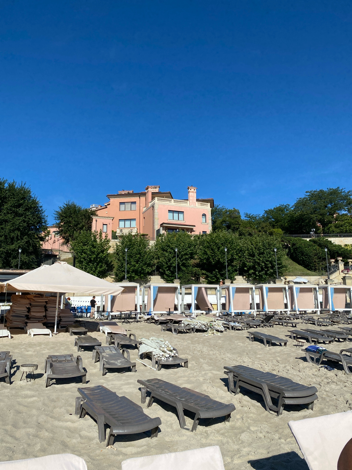 The image captures a sunny day on a sandy beach in Odesa, Ukraine. In the foreground, numerous empty, dark gray plastic lounge chairs are scattered across the sand, some stacked, others individual. Behind the lounge chairs, a row of cabanas with light brown canopies and white frames stretches across the scene. Two people, a man and a woman, are visible near a large, light-colored umbrella in the middle left of the image, appearing to be setting up or organizing. Further back, a large, pale pink building with multiple windows and balconies sits on a hill covered with lush green trees. The sky above is a clear, vibrant blue.