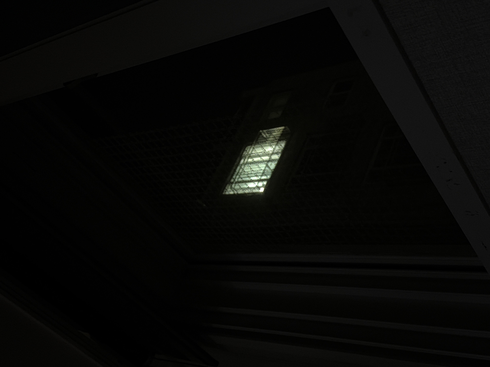 The image shows a nighttime view through a window. The window has a metal grate on the exterior. Inside, a dimly lit building with multiple windows is visible. One section of the building has brightly lit windows, appearing to be offices or rooms. The area outside the window is dark, with some vertical structures that appear to be part of a building or fence. The image was taken in Dongducheon-si, South Korea.