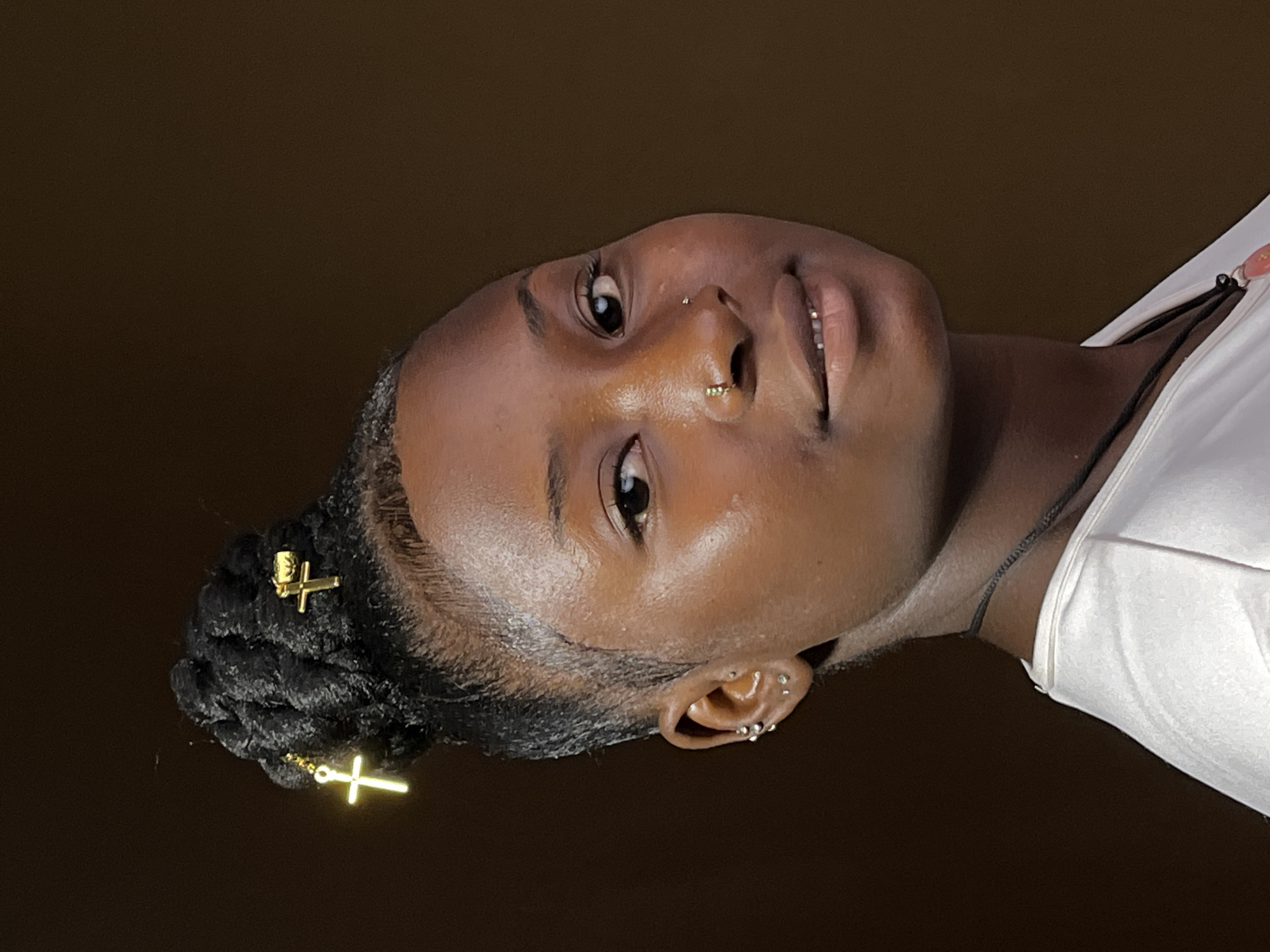 The media file is a close-up portrait of a young person, presented rotated 90 degrees clockwise. When viewed upright, the subject, who has dark skin, is centered in the frame, looking directly towards the viewer with a subtle, confident smile.
Their dark, braided hair is styled into a bun on the left side of their head, adorned with two gold-colored cross-shaped hair accessories. The person has multiple piercings, including a small stud near the bridge of their nose and a gold-colored hoop in their left nostril. Their left ear is visible, featuring several small silver-colored earrings. Their eyes are open and engaged, and the skin exhibits a subtle sheen, catching the light.
The subject is wearing a white garment, likely a t-shirt or tank top, visible on the right side of the frame, along with a thin, dark cord necklace. The background is a plain, dark brown, suggesting an indoor setting or a studio backdrop. There is no visible text in the image. The overall impression is one of composure and directness. The provided location context for the image is Barakin Akawo, Nigeria.