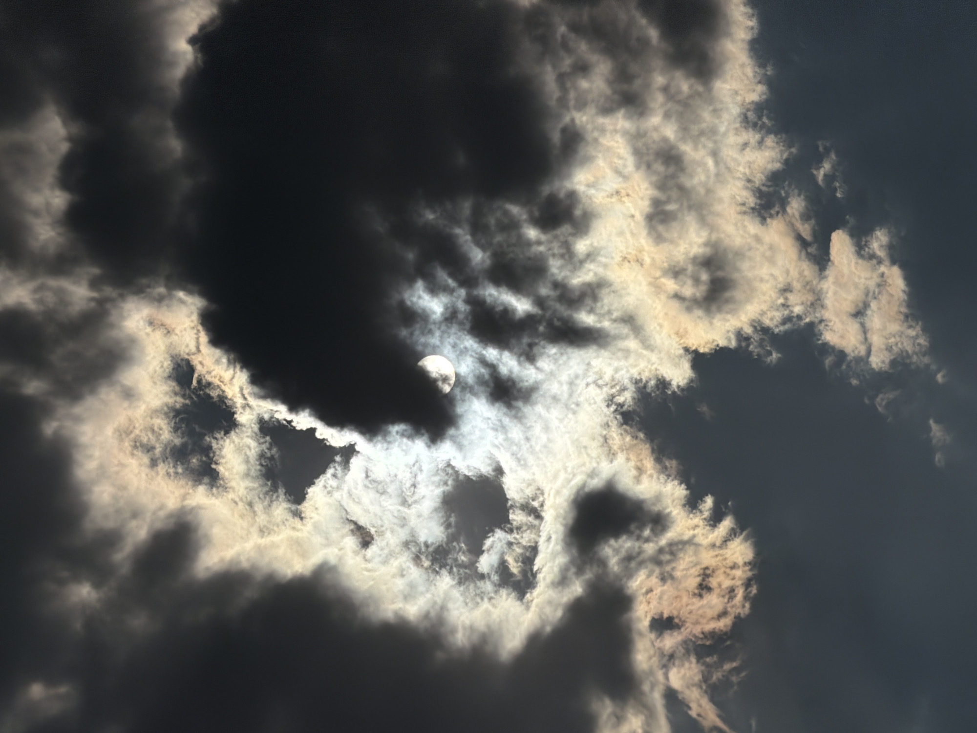 The media file shows a dramatic sky filled with dark, heavy clouds partially obscuring a bright, glowing orb, possibly the sun or moon, near the upper left of the frame. The clouds are a mix of deep grays and blacks, with some edges illuminated by the bright light source, creating a stark contrast. Wispy, lighter clouds are also present, catching the light and adding texture to the scene. The overall impression is one of a turbulent or stormy sky, with the light struggling to break through. There are no people or man-made objects visible, and no text is present. The scene is purely atmospheric and suggestive of a specific weather condition. The location context is Chirchik, Uzbekistan, which could imply the type of cloud formations or lighting conditions common to that region. The time of day is ambiguous, but the strong contrast between light and shadow might suggest either dawn, dusk, or a day with heavy cloud cover. The mood is somewhat ominous due to the dark clouds, yet also awe-inspiring due to the powerful light source piercing through.