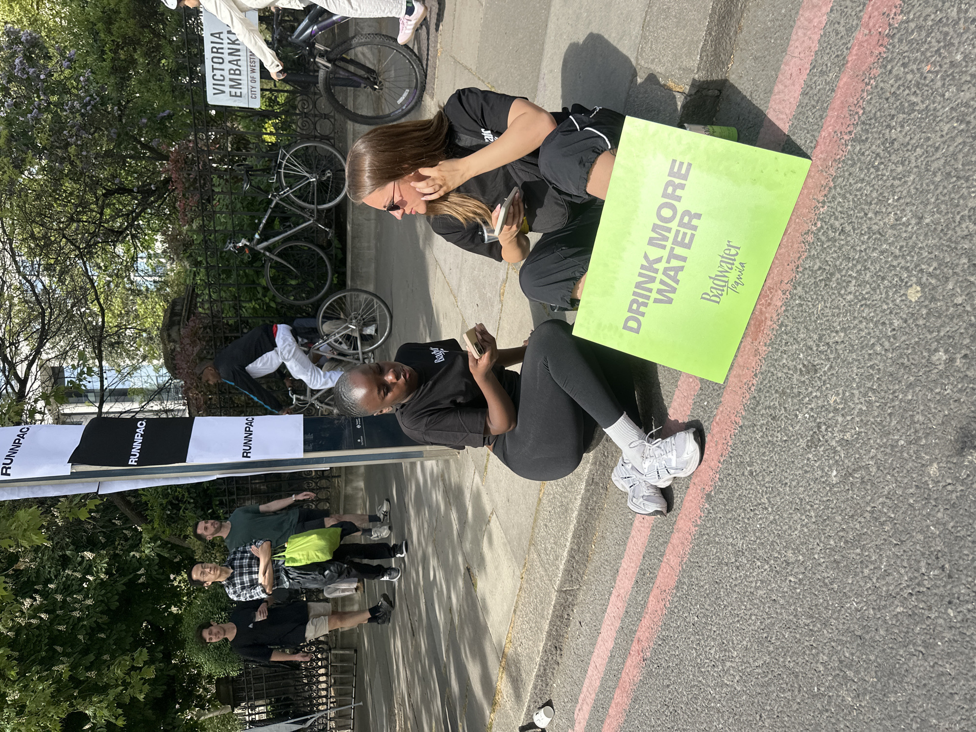 Here is a description of the image:
The photograph captures a scene in the City of Westminster, London, United Kingdom, specifically at Victoria Embankment. Two women are sitting on the curb. One is light-skinned and is wearing sunglasses and is holding a smartphone in her right hand. The other woman is dark-skinned and is also holding a smartphone in her right hand. Next to them on the right side of the photograph is a neon green sign with the phrase, “DRINK MORE WATER Badwater Tequila.” Along the road behind the women are a series of black metal fences where a group of three young males are standing. There is a white banner on top of the metal fences with the text “RUNNPAC.” Behind the fences are multiple bicycles. A Victoria Embankment street sign is visible.