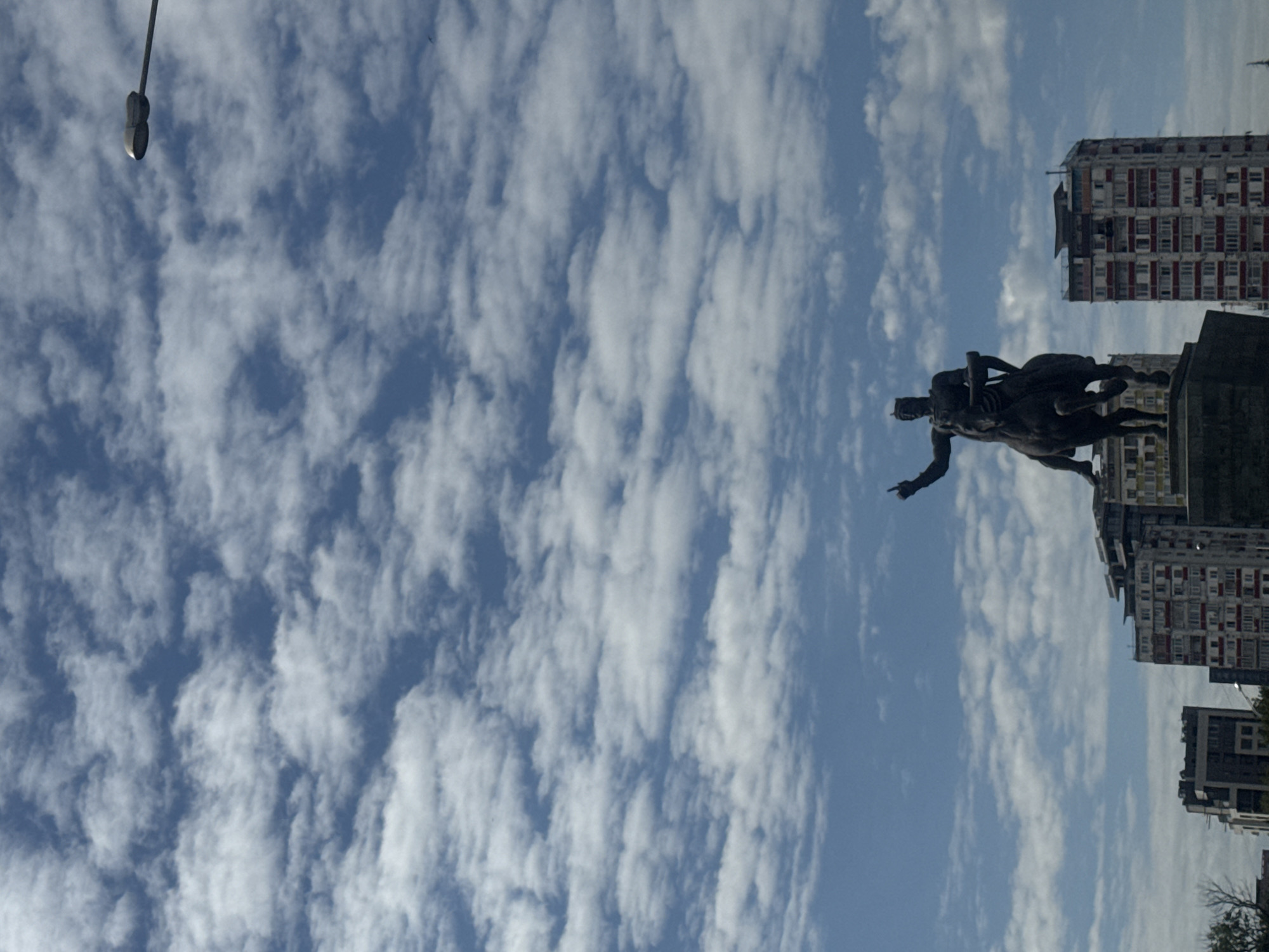The image shows a statue of a man on a horse in Tbilisi, Georgia, with the sky and buildings in the background. The statue is positioned towards the right, with the man pointing forward. Buildings are visible to the right of the statue, and a lamp post is on the upper-left. The sky is filled with white, billowy clouds.