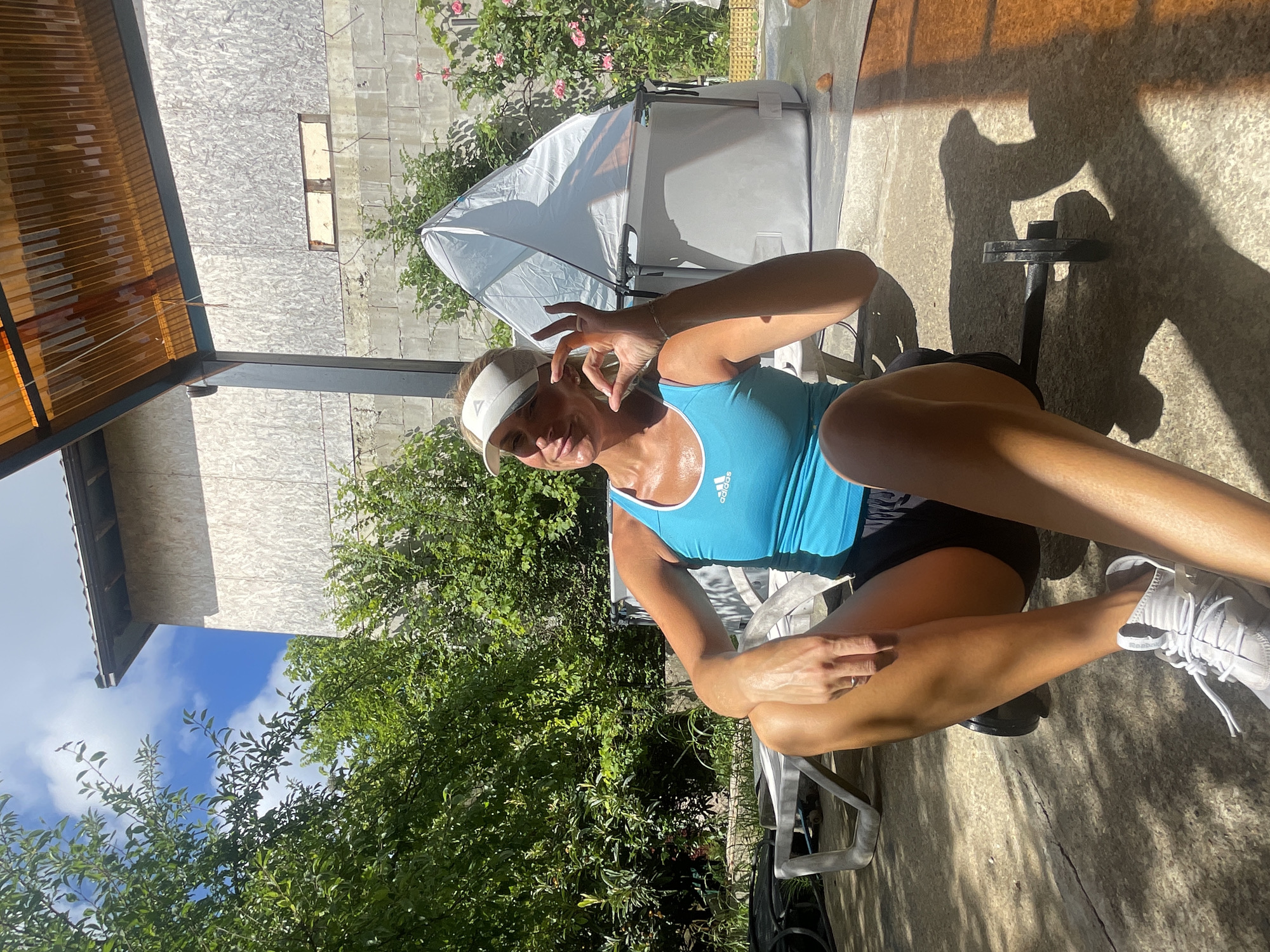 A woman in athletic wear is sitting outdoors in what appears to be a backyard or patio area, possibly in Baranovka, Russia. She is wearing a blue tank top with an Adidas logo, black shorts, a white visor, and grey athletic shoes. The woman is smiling and making an "okay" gesture with her right hand. In the background, there are trees, a white inflatable structure, a building with a wood-paneled exterior, and a corrugated plastic roof structure. A small dumbbell is visible on the ground in front of her. The scene is illuminated by sunlight, casting shadows on the ground.