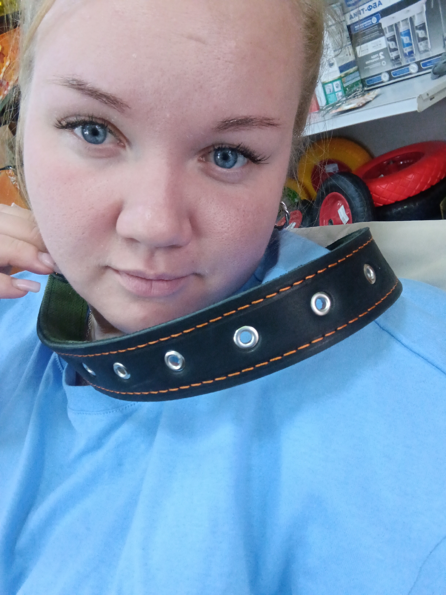 A fair-skinned woman with light blonde hair pulled back in a ponytail poses for a selfie. She has bright blue eyes and is wearing false eyelashes. Her face is softly illuminated, and she has a subtle, thoughtful expression. The woman is wearing a light blue t-shirt. Around her neck, she is wearing a black leather collar with metal grommets and orange stitching.
In the background, there is a shelf with various items, including tires and packaged goods with labels in Russian, suggesting a location in or near Russia. The signage appears to indicate "Анти-Фза" which when translated from Russian would be "Anti-Fza". However, it is unlikely that this is the location in the city of Фоки, Russia because the city does not exist.
