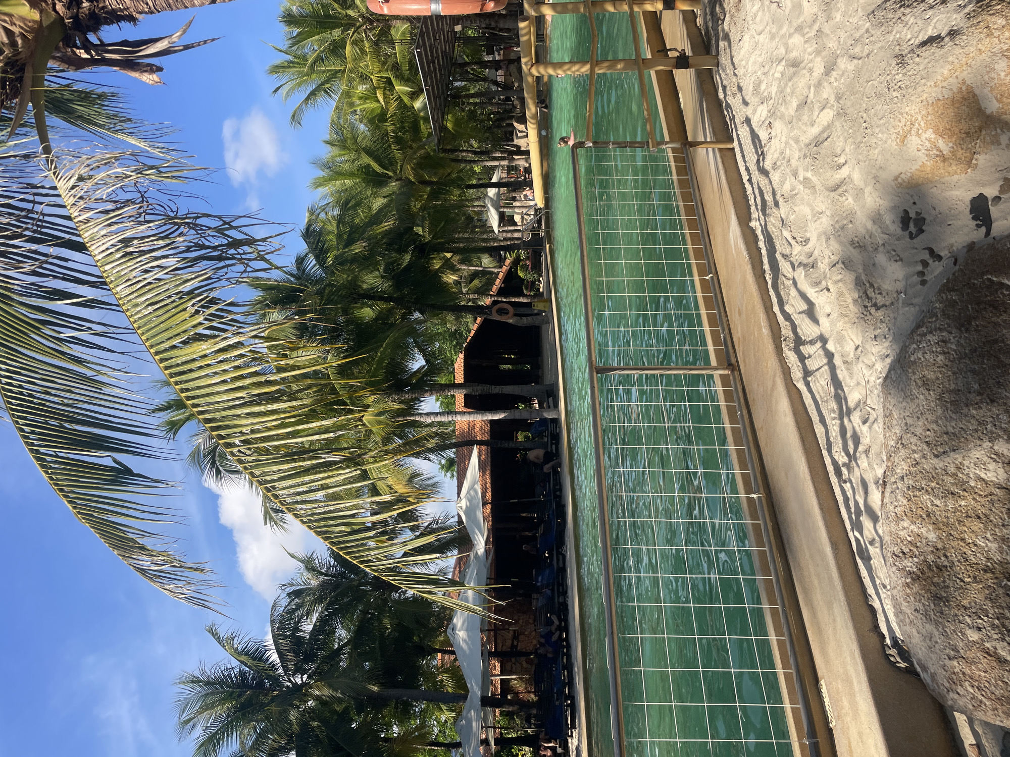 The image depicts a scene at a resort or hotel in Nha Trang, Vietnam, situated near a beach or waterfront. The foreground features a rectangular water feature, possibly a shallow pool or decorative canal, with a grid pattern on the bottom and railings along its edge. Adjacent to the water feature is a sandy area, possibly a beach.
In the background, there are several palm trees, suggesting a tropical or coastal environment. There's also architectural elements suggestive of a resort or hotel structure, including parts of a building with a red-tiled roof, and white umbrellas. There are also lounge chairs.
The scene is captured in daylight. The sky is blue with some white clouds.