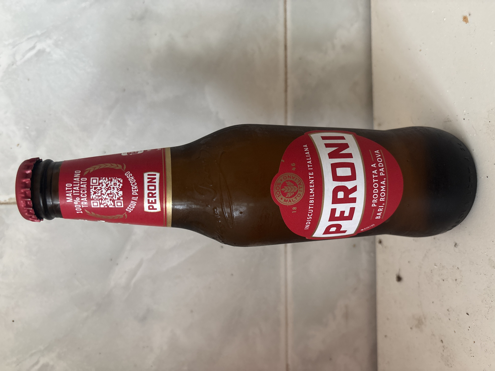 A brown glass bottle of Peroni beer stands upright on a light-colored surface. The bottle has a red cap and a red label with white lettering. The label features the brand name "PERONI" prominently, along with text that reads "MALTO 100% ITALIANO TRACCIATO," "SEGUI IL PERCORSO," and "INDISCUTIBILMENTE ITALIANA." Below the brand name, it also says "PRODOTTA A BARI, ROMA, PADOVA." A QR code is visible on the upper part of the label. The background is a light-colored, textured wall, possibly tiled. The scene is illuminated by overhead lighting. The image was taken in Campomarino, Italy.