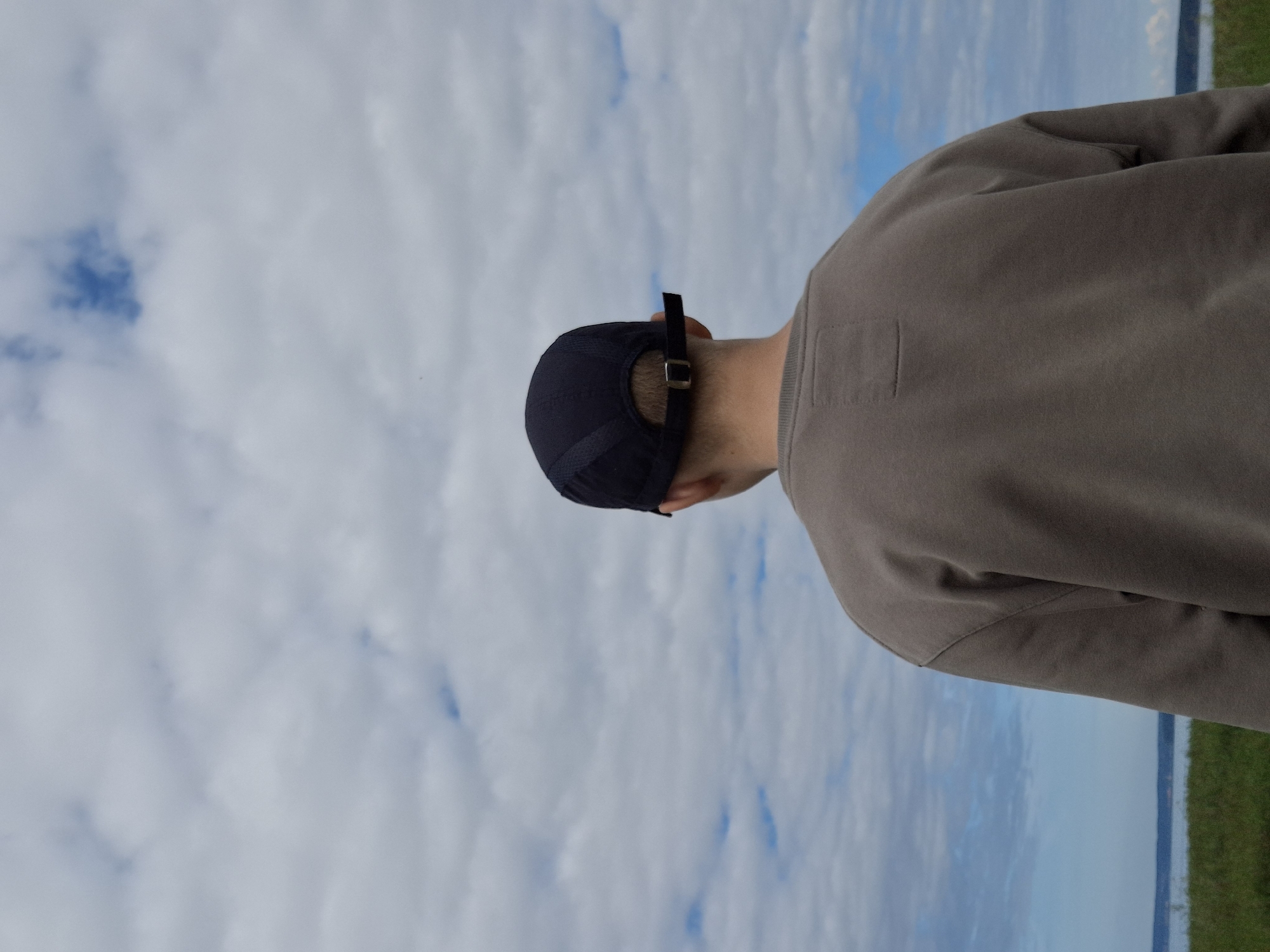 The image, taken outdoors, shows the back of a person wearing a dark baseball cap and a taupe sweatshirt. The person is facing away from the camera, looking towards a cloudy sky and a body of water in the distance, separated by a strip of green grass. The location is identified as Plishkino, Russia. The scene is serene, with no other people or objects prominently featured. The sky is filled with scattered clouds, some of which are dark and dense, while others are lighter and wispy, allowing glimpses of blue sky. The water appears calm, with a faint horizon line visible. The grassy area is somewhat uneven, suggesting a natural landscape. The overall impression is one of quiet contemplation or observation of nature.