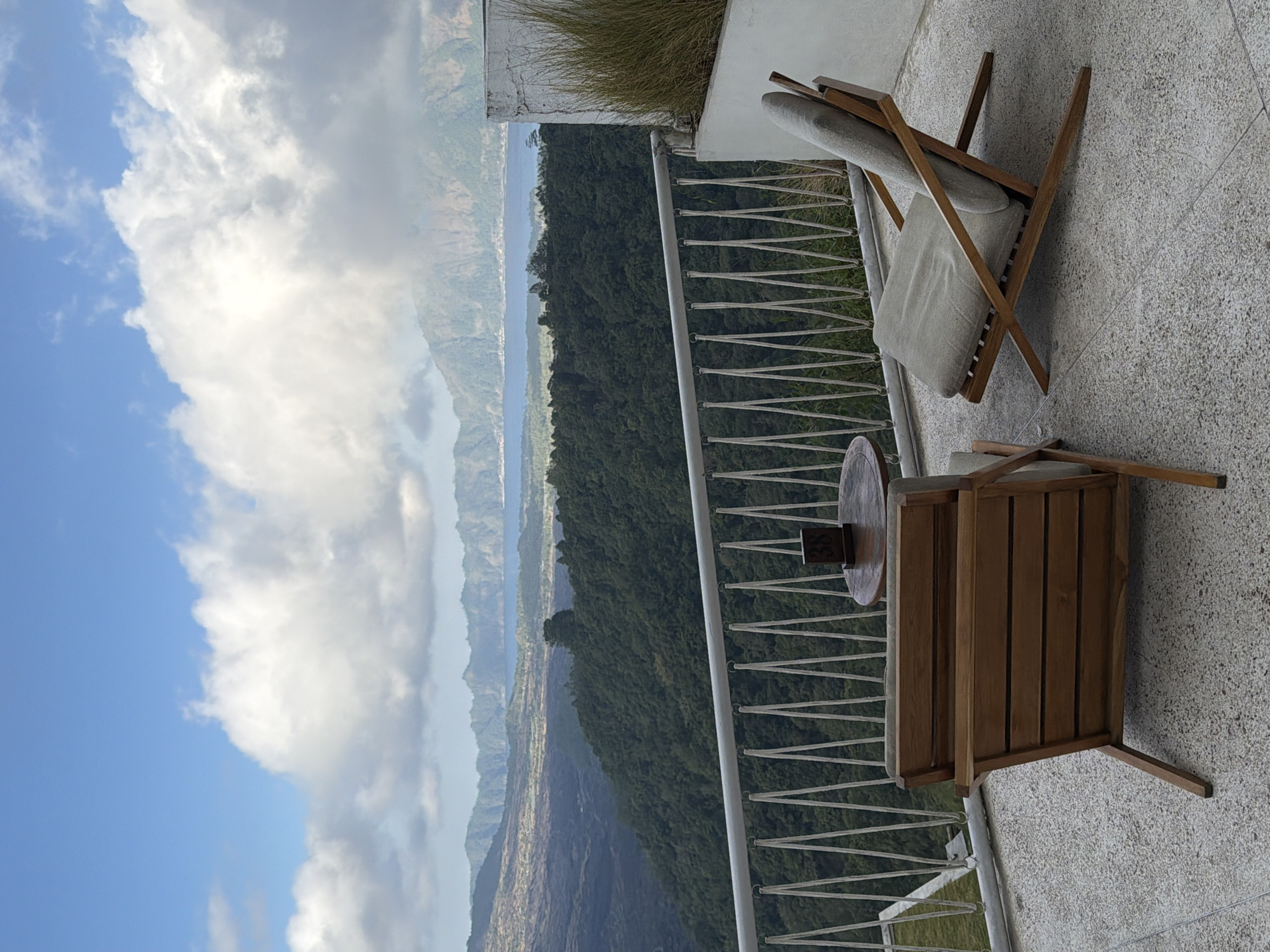 An outdoor seating area in Kintamani, Indonesia, overlooks a vast, verdant landscape. Two modern wooden lounge chairs, upholstered in light gray fabric, are positioned on a textured concrete patio. A small, circular wooden table sits between them, topped with a dark wooden table number displaying "38". Beyond the patio, a white metal railing with rope detailing separates the seating area from the precipitous drop. The background reveals a dense, green forest that cascades down a mountainside. Further in the distance, a serene lake is visible, with layers of misty mountains beyond, their peaks softened by the atmospheric haze. The sky above is a vibrant blue, punctuated by large, fluffy white clouds that cast shadows on the terrain. The image captures a tranquil moment with a breathtaking natural vista.