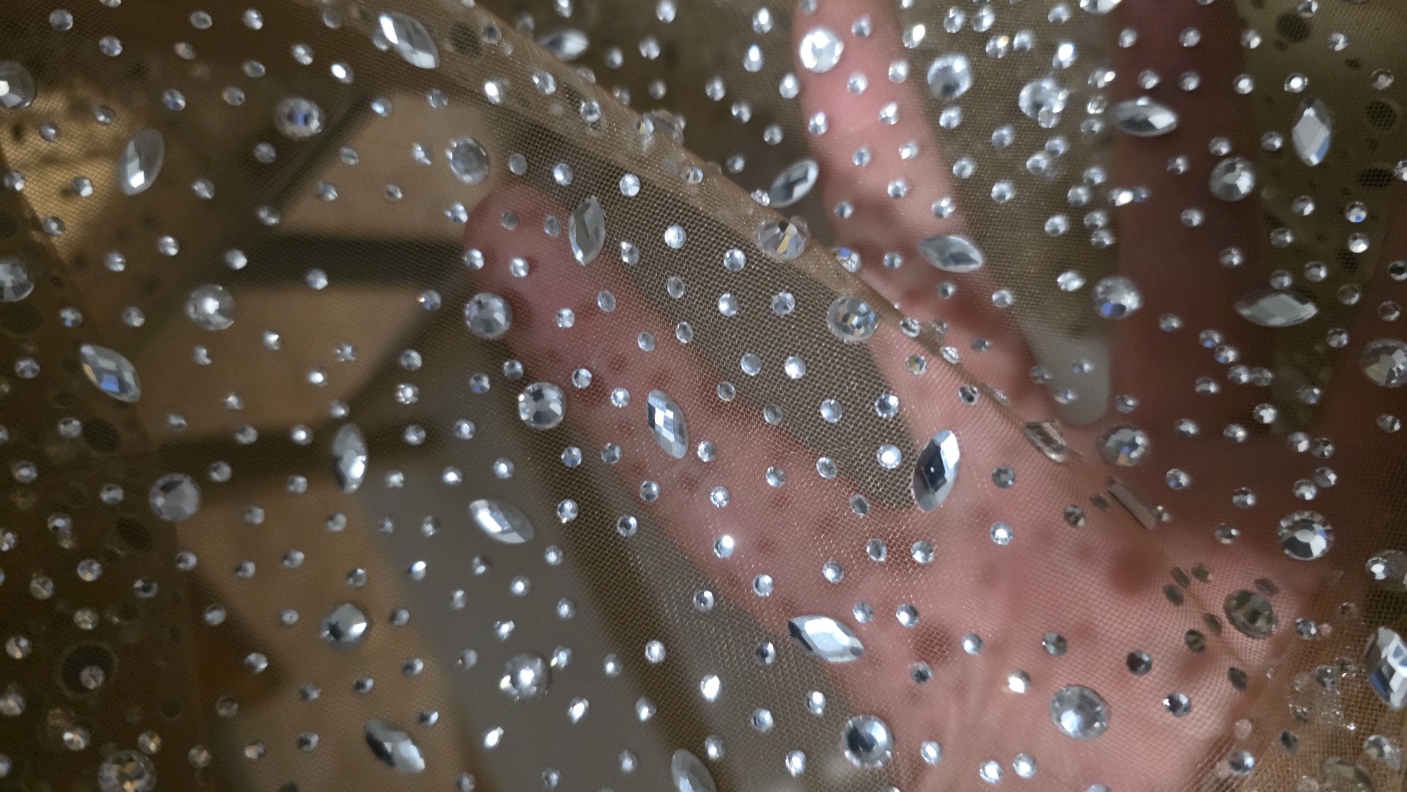 The image presents a close-up view of a sheer, light brown or golden-hued mesh fabric, densely embellished with numerous clear, faceted rhinestones. These reflective adornments vary in size and shape; while most are small and round, several larger, elongated or marquise-shaped crystals are also visible, scattered across the material.
Behind the translucent fabric, a human hand is distinctly visible, its fingers gently pressing against or supporting the material, which highlights the fabric's transparency. The skin tone of the hand is light to medium. The background beyond the hand is out of focus and indistinct, preventing the identification of specific environmental details, though it suggests an indoor setting.
The rhinestones brilliantly catch and reflect light, creating a sparkling and shimmering effect across the entire surface. There is no visible text within the image. The scene emphasizes the intricate texture and decorative quality of the embellished fabric.