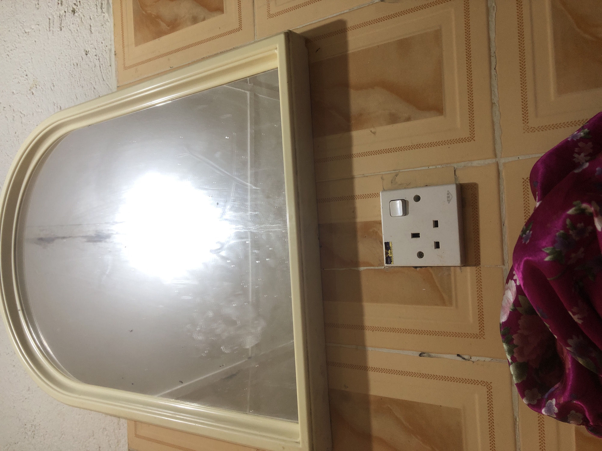 A rectangular electrical outlet with a single switch is mounted on a wall tiled with square, beige tiles featuring a decorative border. The outlet is white and shows signs of wear. Above the outlet, a large, arched mirror with a light-colored frame is mounted on the wall, reflecting a bright light source. Below the outlet, a section of a pink fabric with floral patterns is visible. The wall above the mirror appears to be textured concrete. The overall environment suggests an indoor setting, possibly a bathroom or dressing area. There is no information to suggest this location is in Kabong, Nigeria.