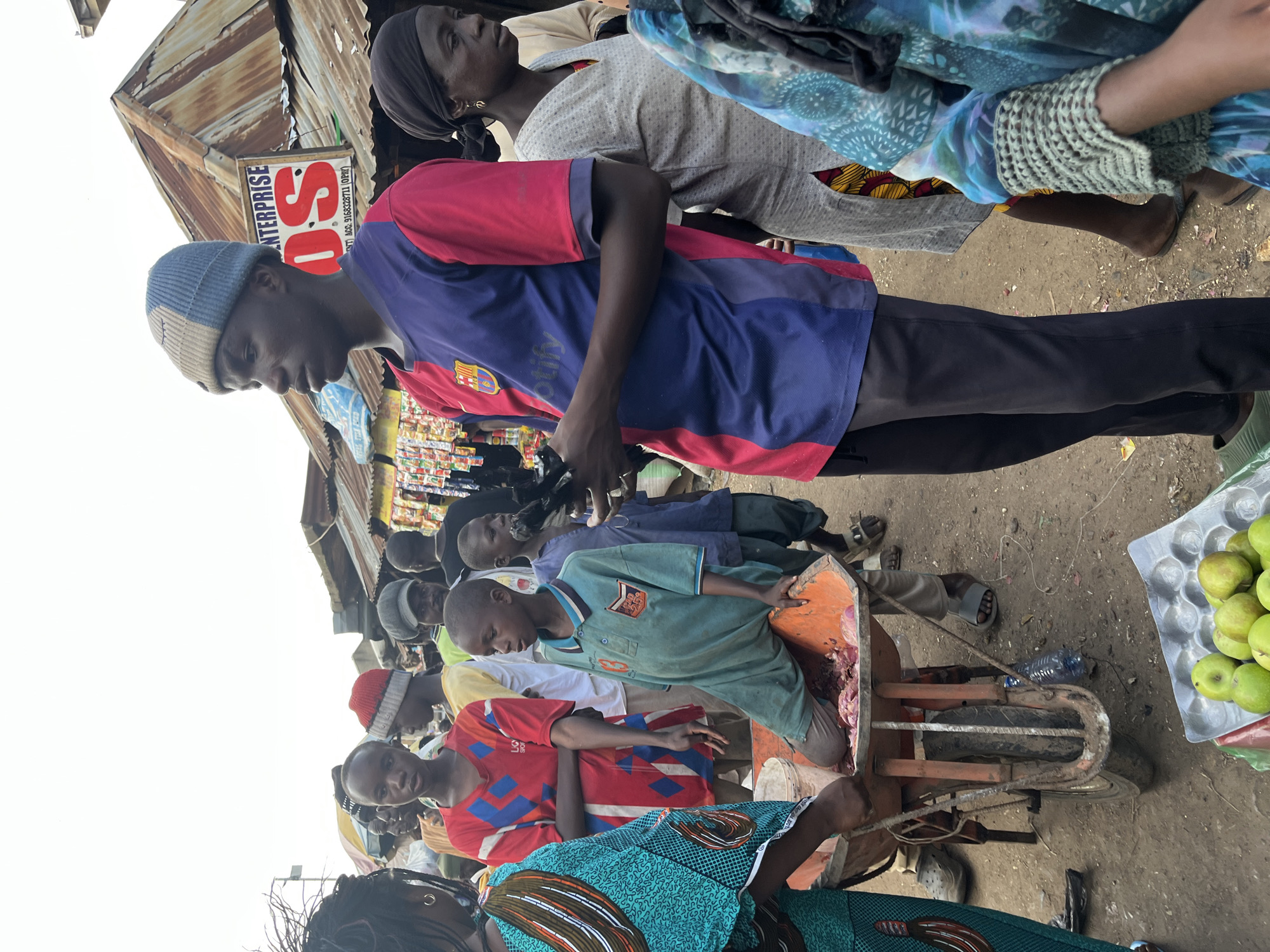 The image depicts a busy outdoor market scene in Rafin Par, Nigeria. In the foreground, a young boy in a teal polo shirt kneels in an orange wheelbarrow filled with raw meat. He is being attended to by a woman in a green and blue patterned dress. To the right of the boy, a young man in a blue and red jersey stands and holds a black object, possibly a small animal, in his hands. He wears a blue and grey knitted beanie. In the background, several other people are visible amidst market stalls with corrugated metal roofs. One stall displays a sign for "ENTERPRISE OS." A tray of green apples sits on the ground in the lower right corner. The ground is dirt and appears to be a well-trafficked area.