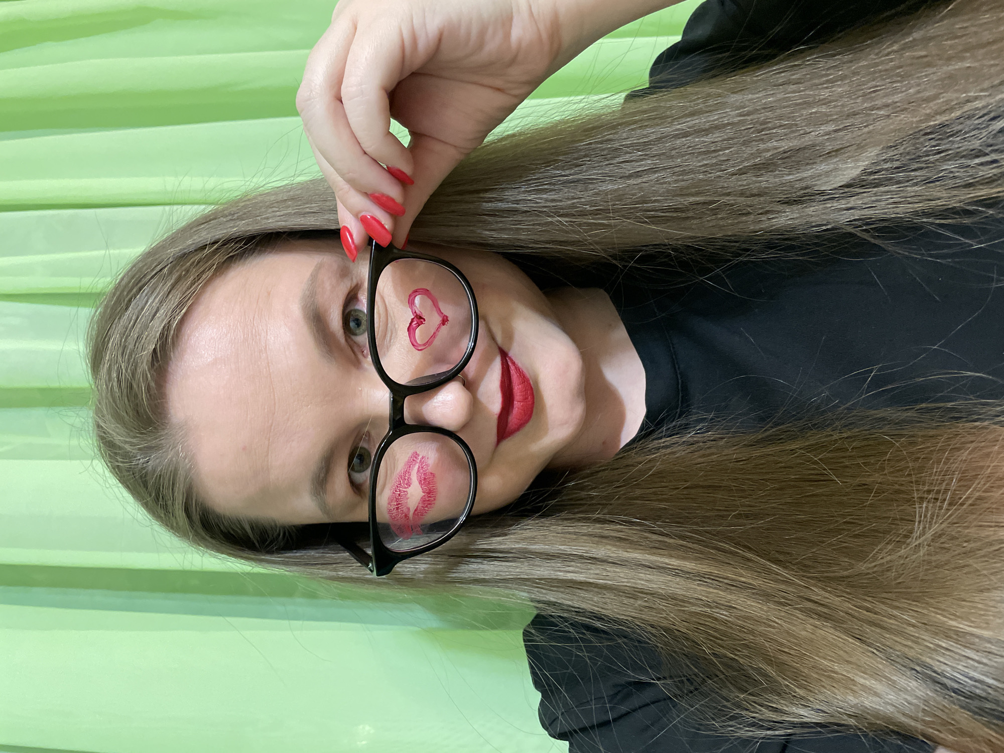 The image is a vertical, close-up portrait of a woman looking directly into the camera, taken in Izhevsk, Russia. She has long, straight brown hair that cascades over her shoulders and is wearing a black top. Her fingernails are painted with bright red polish, and she is also wearing bold red lipstick.
The woman is holding a pair of black-framed glasses up to her face with her right hand. On the left lens of the glasses, a small, hand-drawn red heart shape is visible, positioned over her left eye. On the right lens, there is a clear impression of red lipstick, resembling a kiss print, situated over her right eye. Her expression suggests a playful and affectionate mood, with a slight smile.
The background behind her is a light, vibrant green, appearing to be a textured fabric or surface, possibly a curtain or blind, with visible horizontal folds. The overall impression of the image is one of lightheartedness and charm. There is no visible text in the image.