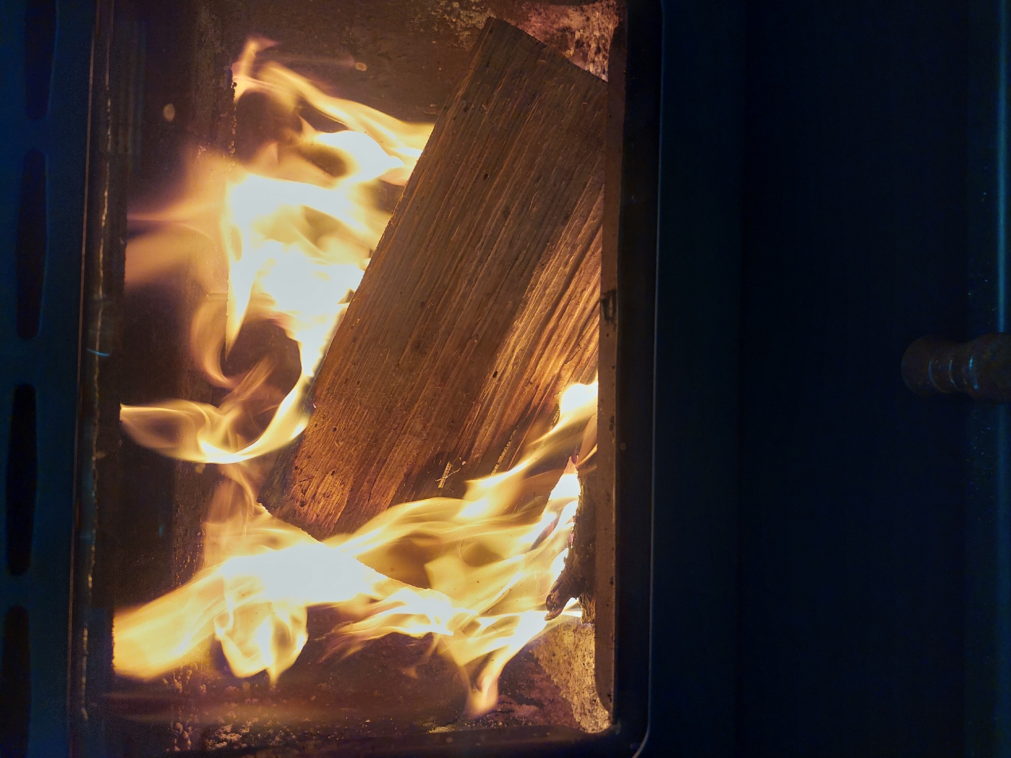 The image depicts a wood-burning stove in operation. A large piece of firewood is positioned within the stove, surrounded by active flames. The interior of the stove appears dark and sooty. The stove door is ajar, revealing the fire. The right side of the image shows the dark blue exterior of the stove. The scene is indoors, likely a residential setting. The location is Nedelino, Bulgaria, though there are no elements in the photograph to confirm the location.