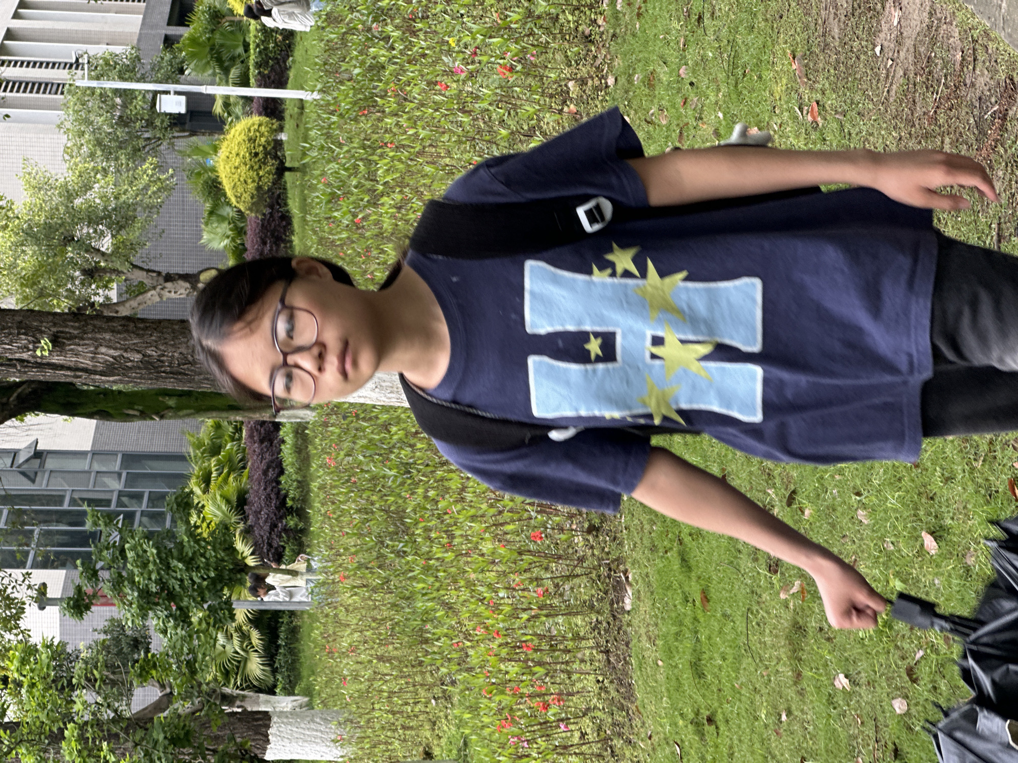 A young person with dark hair and glasses is standing outdoors in a grassy area with trees and shrubs. They are wearing a dark blue t-shirt with a large light blue "H" design featuring yellow stars. The person has a black strap across their shoulder. They are holding a black umbrella in their right hand. In the background, there is a building with windows. The location is likely within Jiangjin, China, based on the provided information. The scene appears to be a casual outdoor setting, possibly a park or garden area.