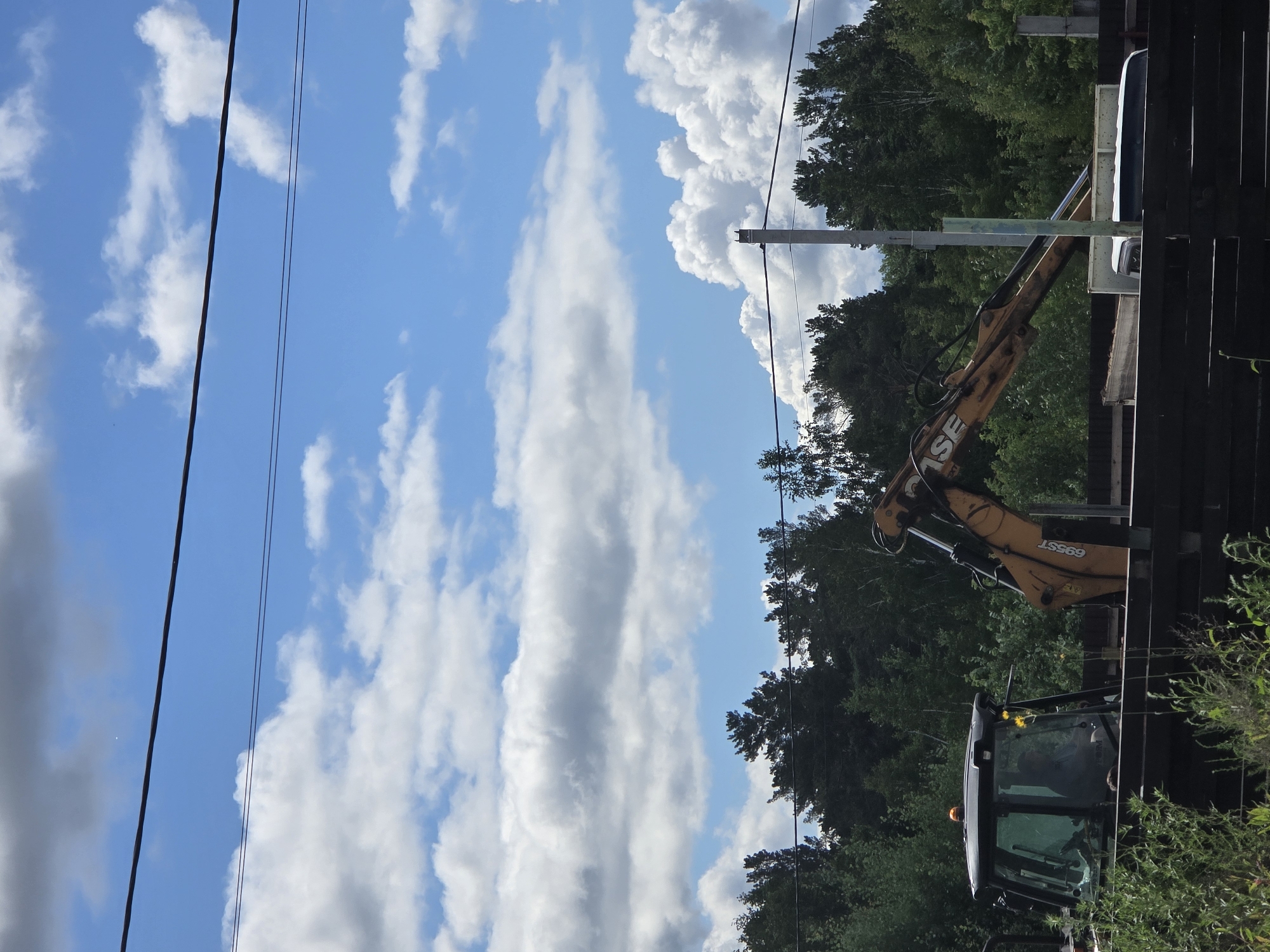 A backhoe loader is parked in an outdoor area with trees and a cloudy blue sky. The backhoe, which is yellow and black, has its arm extended. A person is visible inside the cabin of the backhoe. The area appears to be under construction or renovation, with a wooden fence and some construction materials in the foreground. The location is identified as Ermolaevo, Russia.