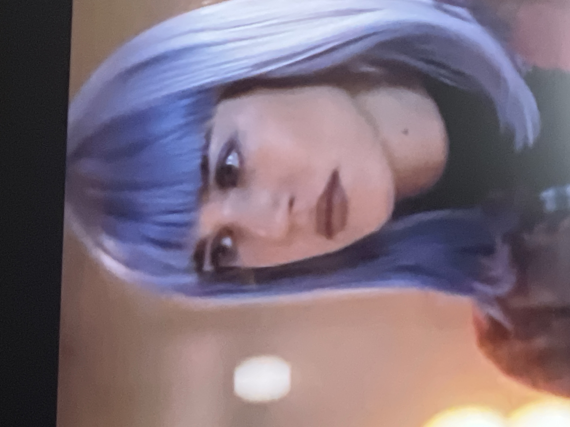 This media file is a close-up, vertically oriented image of a person's face and hair. The subject appears to be female, with light purple or blue hair styled in a bob with bangs. Her face is turned towards the viewer, with her eyes looking directly ahead. She has dark eyebrows, and her lips are painted a reddish-brown color. There is a small, dark mole visible on her cheek. The lighting is somewhat dim, with a soft glow emanating from an unseen source, suggesting an indoor setting. The background is blurred, with warm tones of orange and yellow. The overall mood of the image is somewhat ambiguous due to the angle and lighting, but the direct gaze of the subject could be interpreted as confident or introspective. There is no discernible text in the image. The location context provided is Baošići, Montenegro, but there are no visual cues within the image to confirm this specific location.