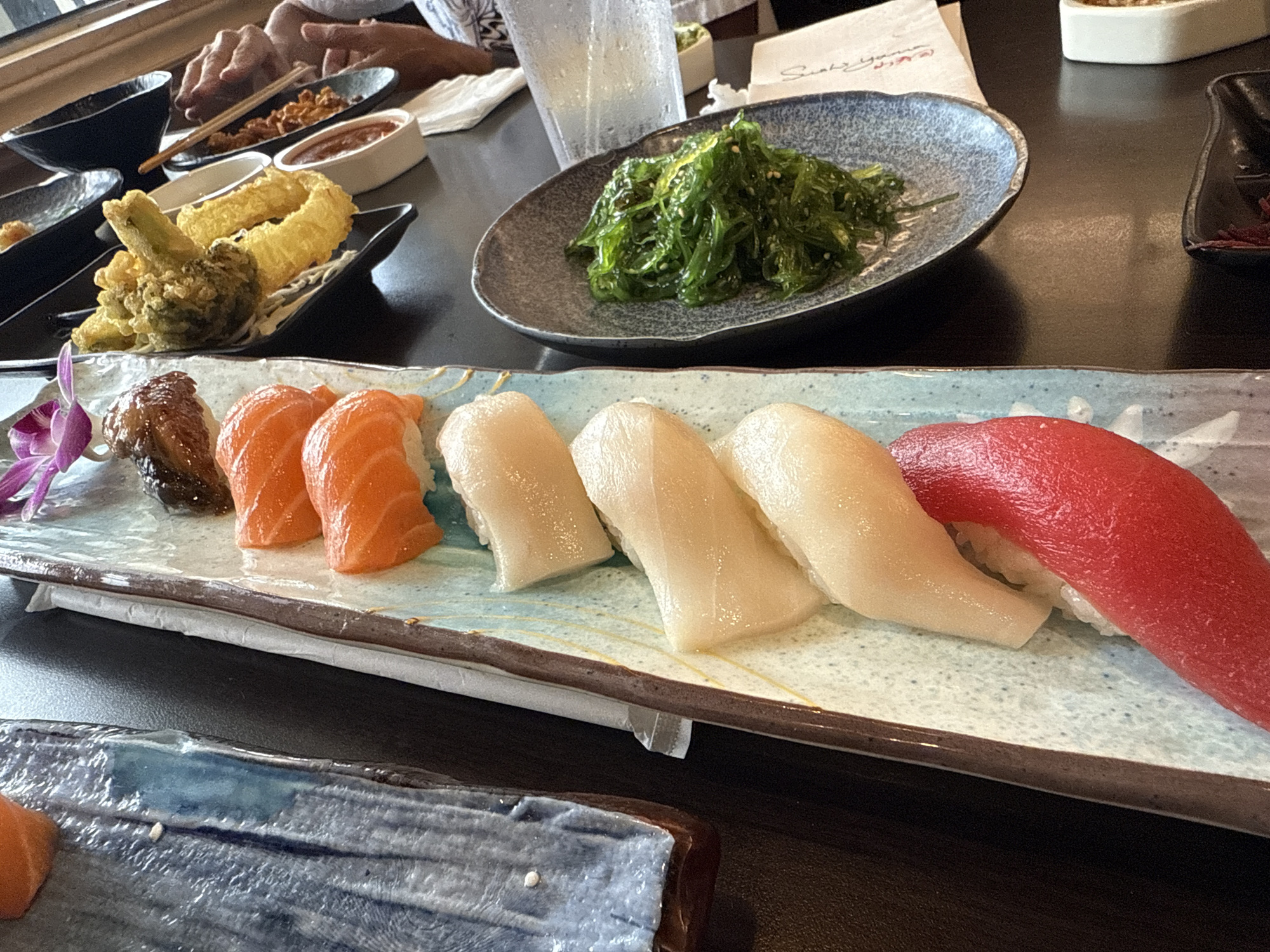 The image shows a table at a restaurant in Orlando, Florida, United States. A variety of Japanese dishes are arranged on the table, including a sushi platter with eel, salmon, white fish, and tuna nigiri. There is also a plate of seaweed salad and a dish of tempura. People are visible in the background.