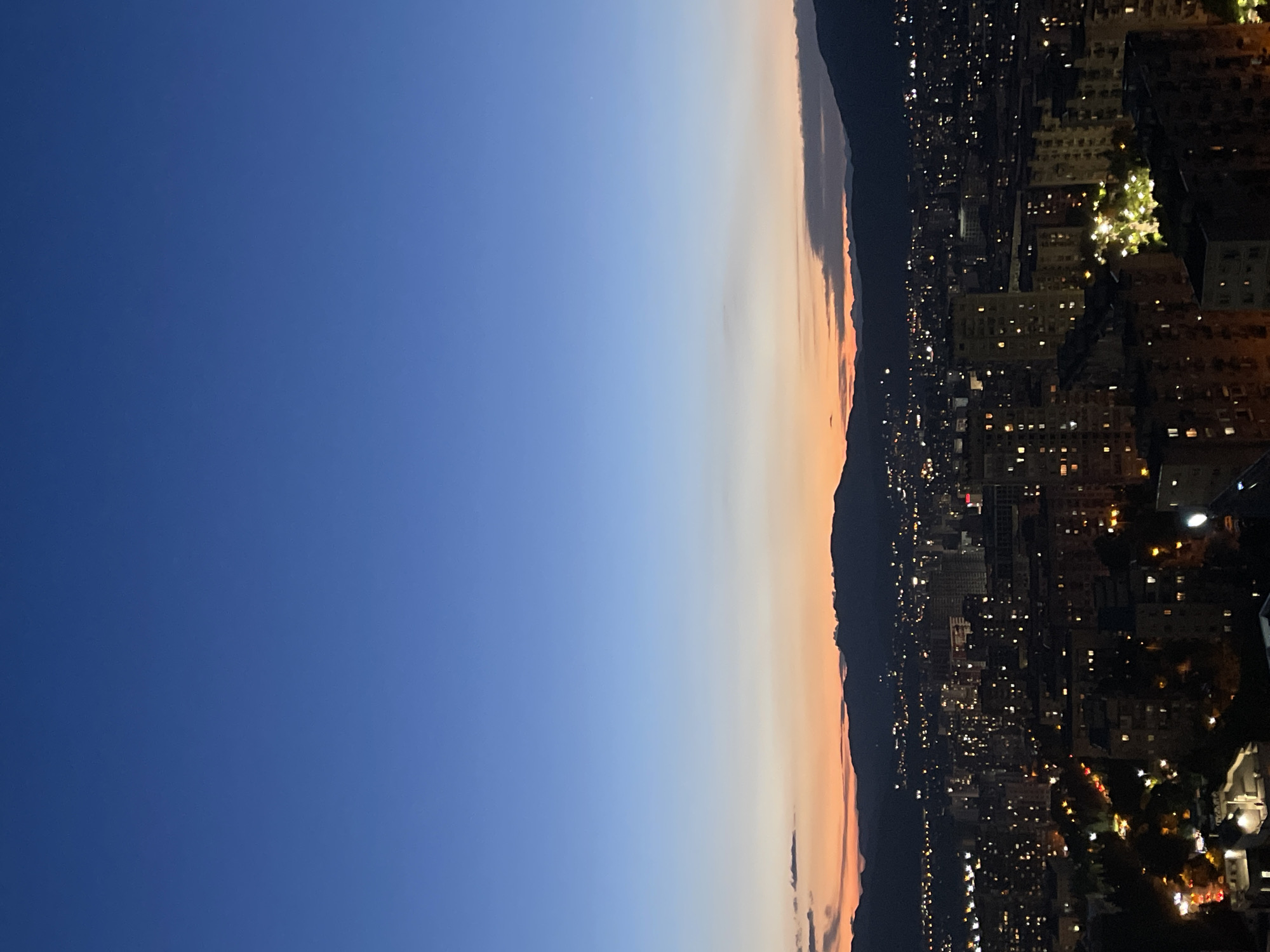 An elevated, wide shot captures the city of Tbilisi, Georgia, at dusk. The sky transitions from a deep indigo at the top to hues of soft blue, orange, and pink near the horizon, creating a serene twilight atmosphere. Silhouetted mountains form a dark backdrop to the urban landscape. Below, the city lights twinkle, illuminating apartment buildings and streets. The image focuses on the sprawling architecture and the gradual emergence of artificial light against the fading natural light. No people are visible in the frame.