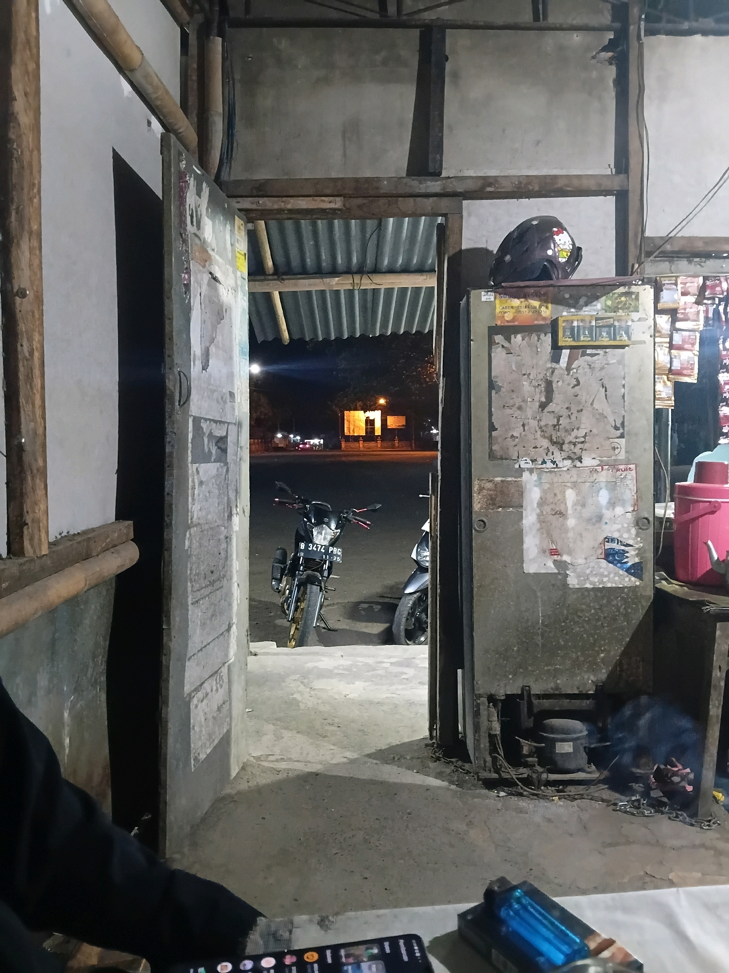 The image depicts an outdoor scene at night in what is described as Bantarbarang, Indonesia. In the foreground, a person's arm, clad in a black long-sleeved garment, is visible on the left, holding a smartphone. On a table in front of the person, a blue lighter rests next to the smartphone.
The midground of the image features an open doorway leading into a dimly lit interior. Through the doorway, a motorcycle with license plate "B 3474 PBG" is parked on the right. Another motorcycle is partially visible behind it.
To the right of the doorway stands a large, weathered metal object, possibly a refrigerator or a large cabinet, covered with peeling posters and stickers. On top of this object, a dark helmet with a Hello Kitty design is placed. Adjacent to this, to the far right, a bright pink thermos is visible, along with other items on shelves.
The background shows a street scene at night with some structures and distant lights. The overall environment appears to be a casual, possibly commercial or residential area, with rustic construction elements such as wooden beams and corrugated metal roofing visible.