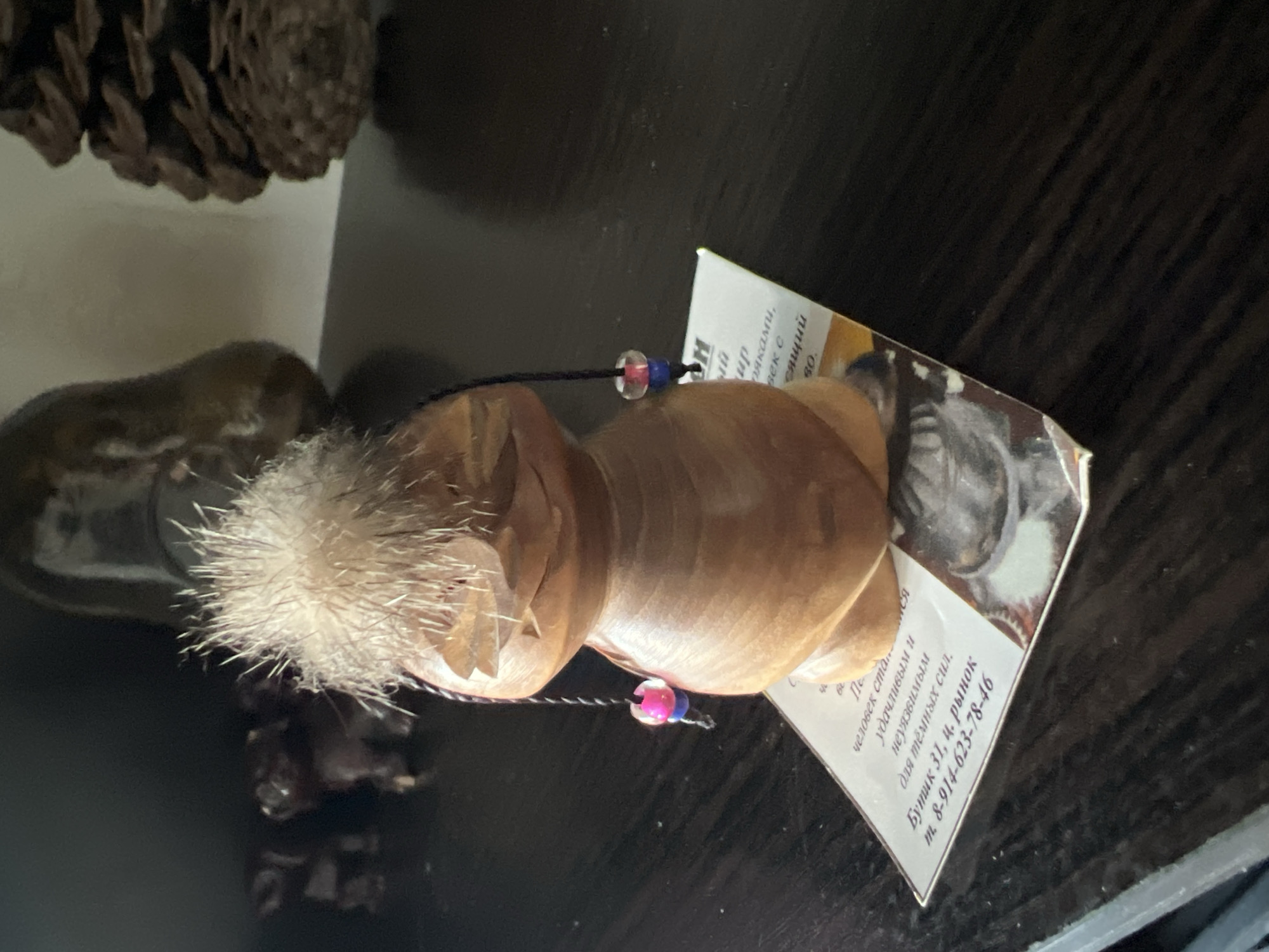 The image shows a small wooden figurine with a tuft of fur on its head, resting on a piece of paper with Russian text. The figurine has beads with pink and blue accents hanging from its sides. In the background, blurred objects, possibly other figurines and a pinecone, are visible on a dark surface. The overall setting appears to be indoors, on a shelf or table. There is no discernible information to place this scene in the city of Dnp Ryzhiki, Russia.