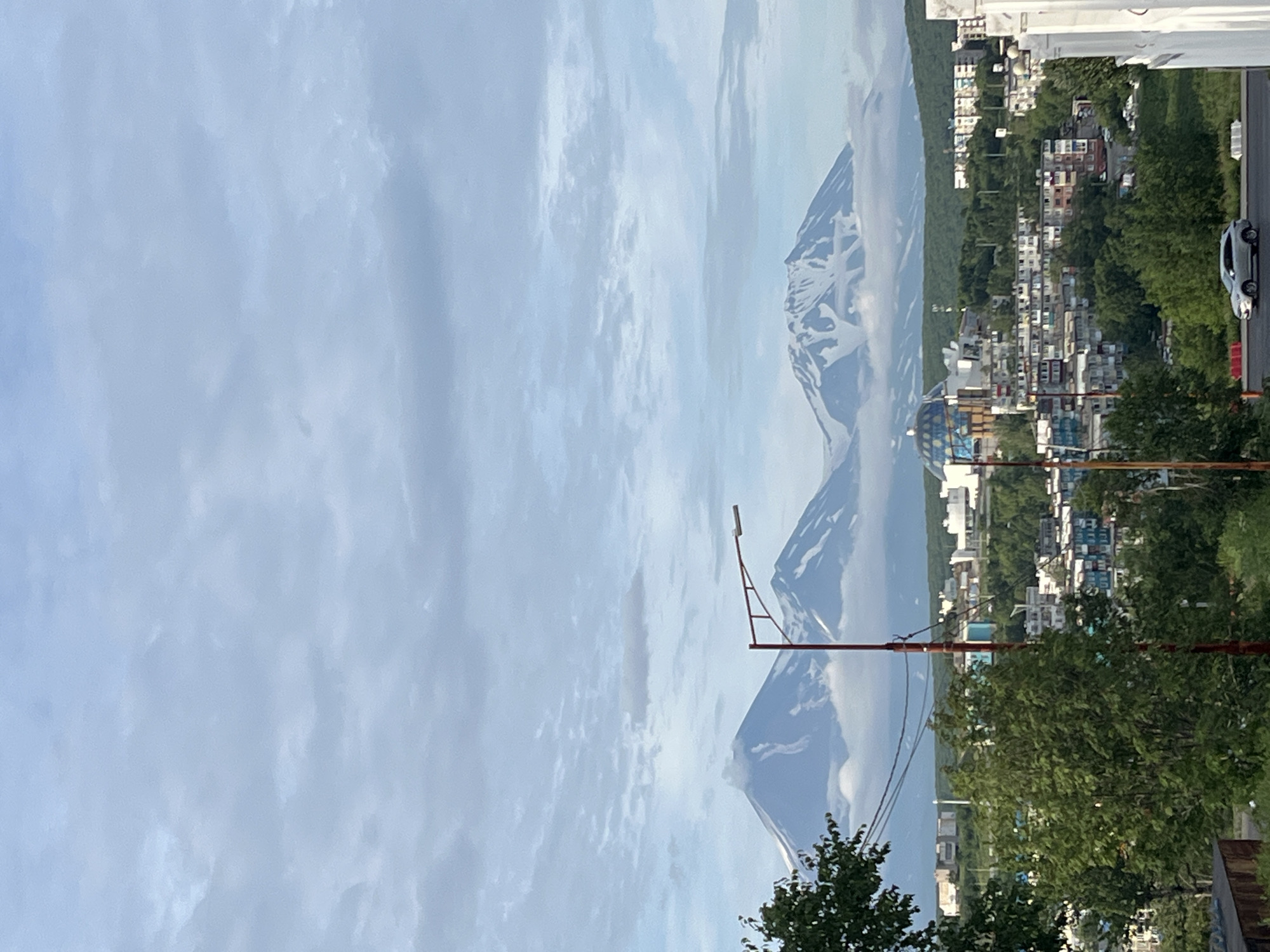 The image depicts a panoramic view of Petropavlovsk-Kamchatsky, Russia, with the city's architecture nestled at the foothills of snow-capped volcanoes under a cloudy sky. In the foreground, a green tree obscures part of the view, and a street light with a red pole stands prominently. The middle ground reveals a cityscape characterized by apartment buildings of varying colors and heights, some with balconies and windows. A distinctive blue-domed building with glass panels is visible amidst the urban landscape. In the background, three imposing volcanoes rise majestically, their peaks covered in snow and partially obscured by clouds. A silver car is seen driving on a road in the lower right portion of the image.