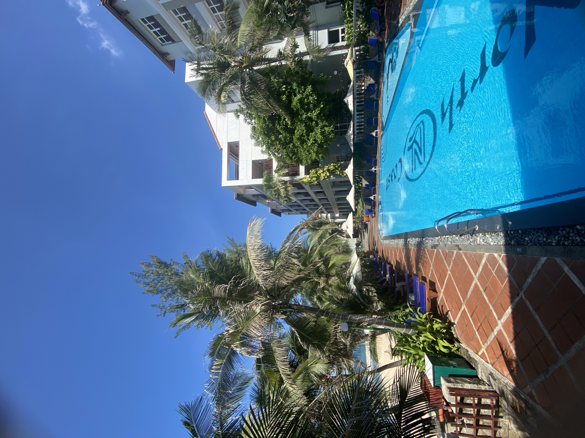 The image depicts a bright, sunny day in Phu Thuy Ward, Vietnam, featuring a resort or hotel swimming pool area. The foreground is dominated by the vibrant blue water of a large swimming pool, with a dark, stylized logo visible on the pool floor. The pool area is paved with terracotta tiles arranged in a diamond pattern. To the left of the pool, a pathway is lined with blue lounge chairs and white umbrellas. Beyond the lounge chairs, lush green palm trees and other tropical foliage are visible, with the sandy beach and ocean in the background. Several resort buildings with multiple stories and balconies are visible behind the trees and pool area. The sky is a clear, brilliant blue with no clouds. The scene suggests a tranquil and luxurious vacation setting.