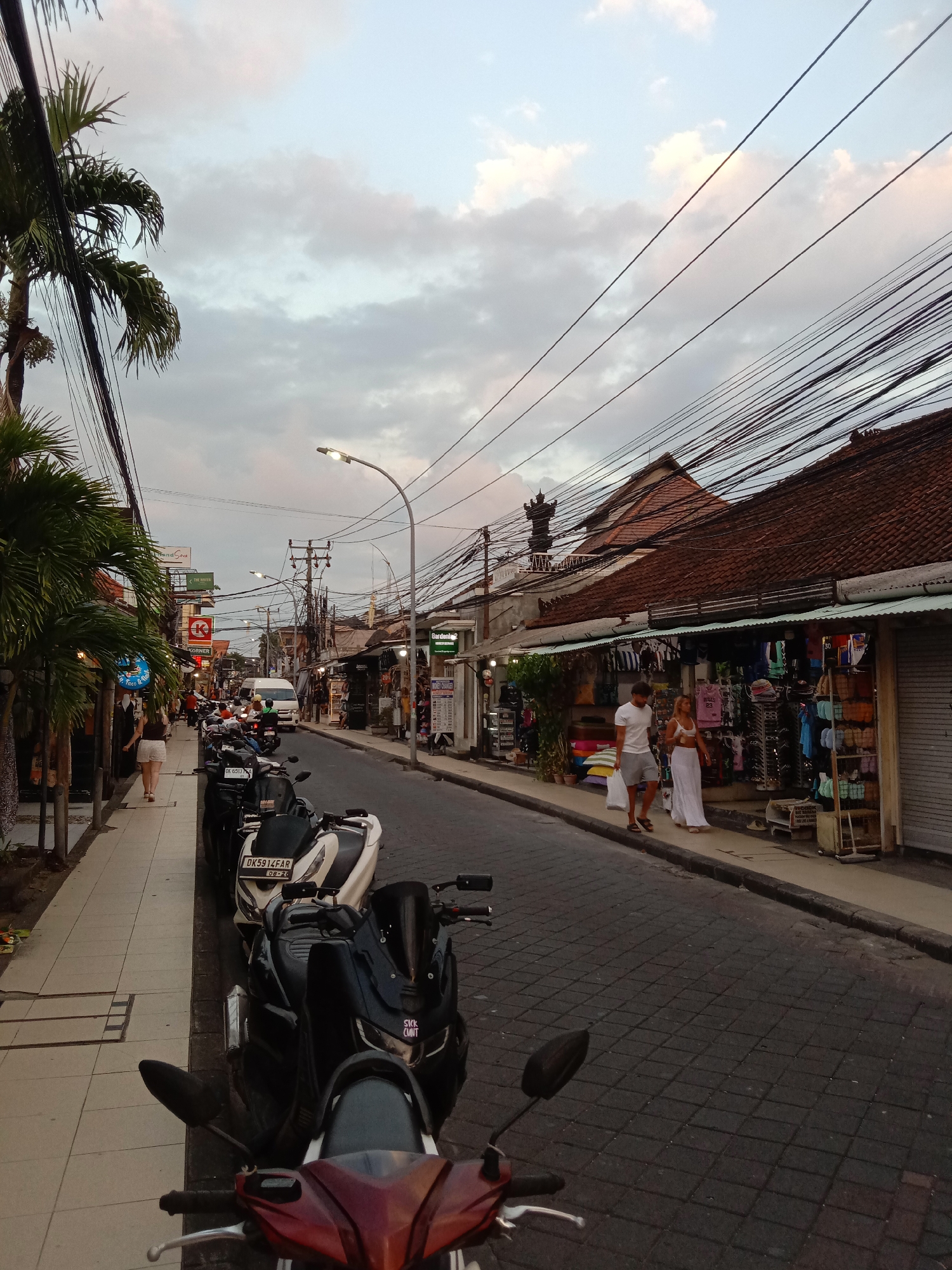 The image depicts a street scene in Kuta, Indonesia, at dusk. A narrow road, paved with dark interlocking stones, is lined with shops and palm trees. Numerous motorcycles are parked along the sidewalk on the left side of the road. On the right side, two people, a man and a woman, walk away from the viewer, passing by a shop displaying clothing and souvenirs. The sky is cloudy with hints of orange and pink, indicating the setting sun. Overhead, a dense network of electrical wires is visible against the sky. The buildings on either side of the street are low-rise with tiled roofs.