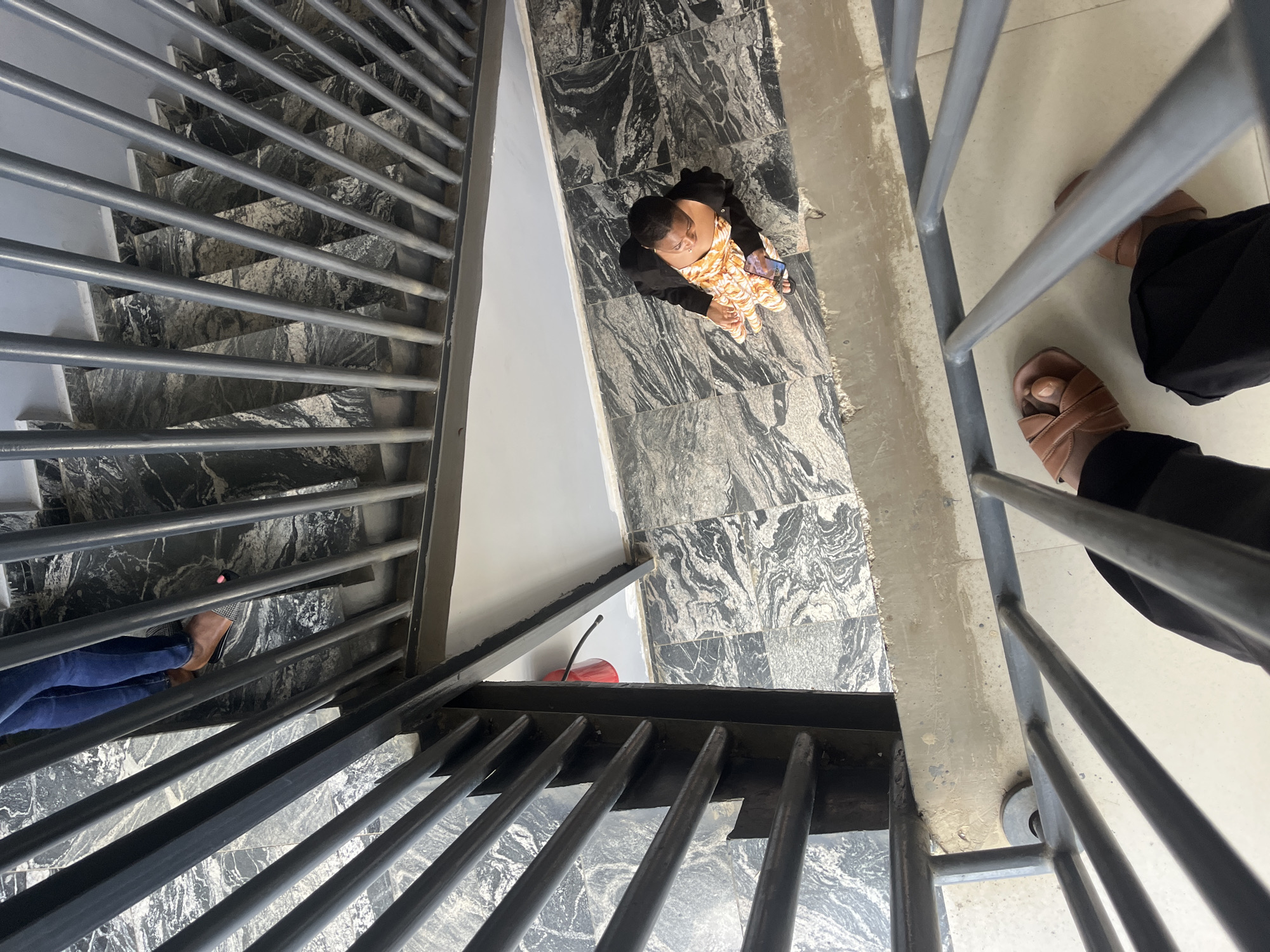 The image shows a person wearing a black jacket and an orange and white patterned dress, holding a smartphone, standing on a tiled floor. The floor is decorated with a marbled pattern. The person is positioned on a landing that appears to be part of a staircase. Metal railings, characteristic of a staircase, are visible in the foreground and extend upwards and downwards. In the background, another person's legs wearing blue jeans and sandals are visible, suggesting they are also on the staircase, possibly descending. The lighting suggests it is daytime. The overall environment appears to be an indoor or semi-outdoor staircase structure. No specific landmarks identifying Karum, Nigeria are visible.