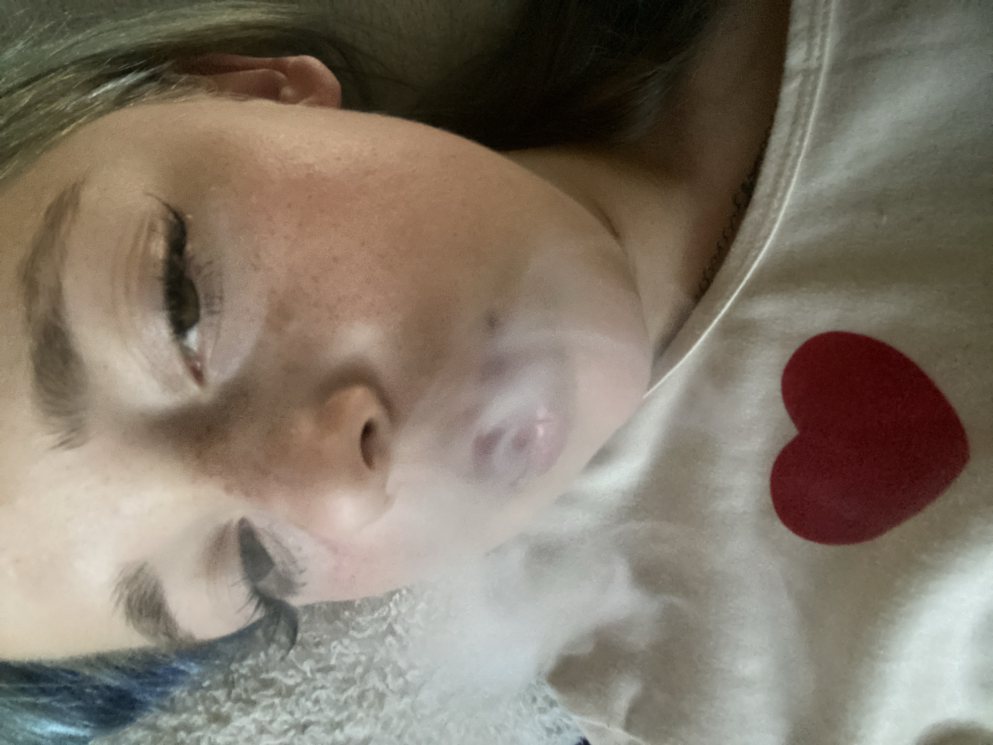 The image is a close-up, head-and-shoulders shot of a young person with light skin, freckles, and light-colored hair, some of which has blue streaks. The person is wearing a white t-shirt with a red heart graphic. They appear to be exhaling a cloud of white vapor, obscuring their mouth and nose. The background is a textured, light-colored surface, possibly a blanket or bedding. The lighting is soft and appears to be coming from above. The image was taken indoors. The location in Nova Praha, Ukraine, is not discernible from the image.