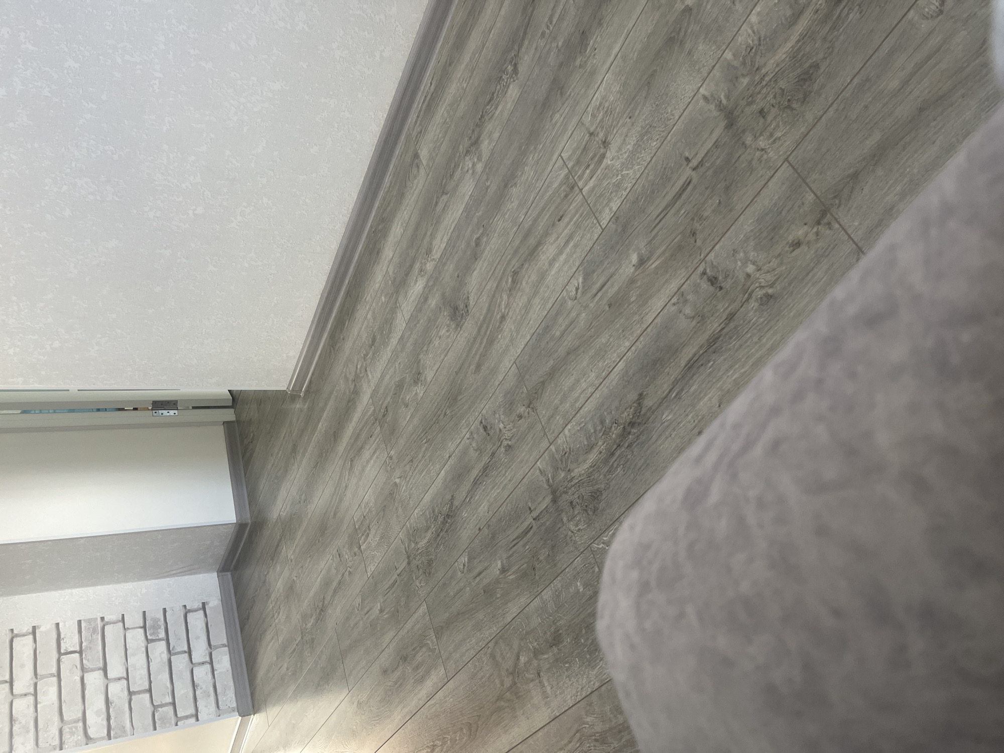 The image shows a room interior with gray wood-patterned flooring, white walls, and a section of brick-patterned paneling at the base of one wall. The flooring appears to be laminate or vinyl plank. A portion of a gray object, possibly a sofa or chair, is visible in the right side of the frame. The scene is otherwise devoid of people or specific objects to identify a location.