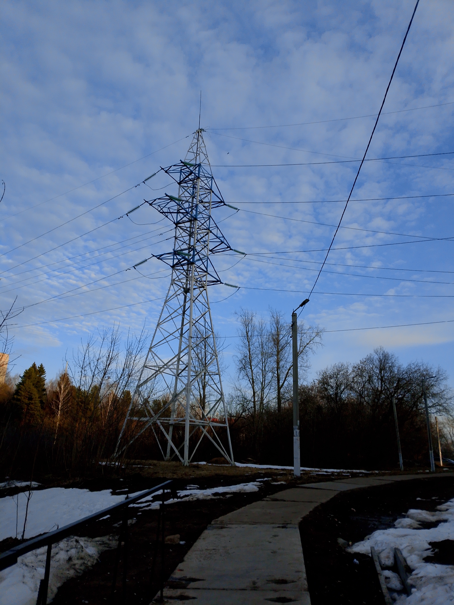 The image depicts an electrical transmission tower in a partially melted, snowy environment. The tower is painted with white and blue paint. Bare trees and vegetation are visible in the background. A paved walkway runs through the scene, with patches of snow and dirt along its edges. A street light stands next to the path. The sky is blue with scattered clouds. Based on the image's visual details and known landmarks, the location is within the city of Izhevsk, Russia.