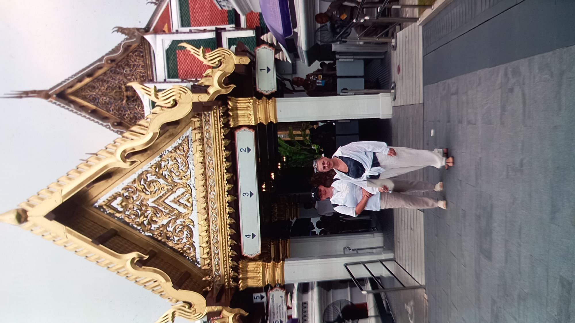 Two women, one wearing a white top and beige pants, the other a white open shirt over a dark tank top and white pants, stand on a paved walkway in front of a building with ornate golden architecture. The building features multiple tiered roofs with intricate golden decorative elements. Above an entryway, signs with numbers 4, 3, and 2 are visible, with arrows pointing downwards. To the right of the women, a sign with the number 1 and a downward arrow is also present. Security personnel in uniform are visible in the background near the entrance. The overall setting appears to be a temple or a significant cultural site in a tropical climate, likely in Thailand given the architectural style.
