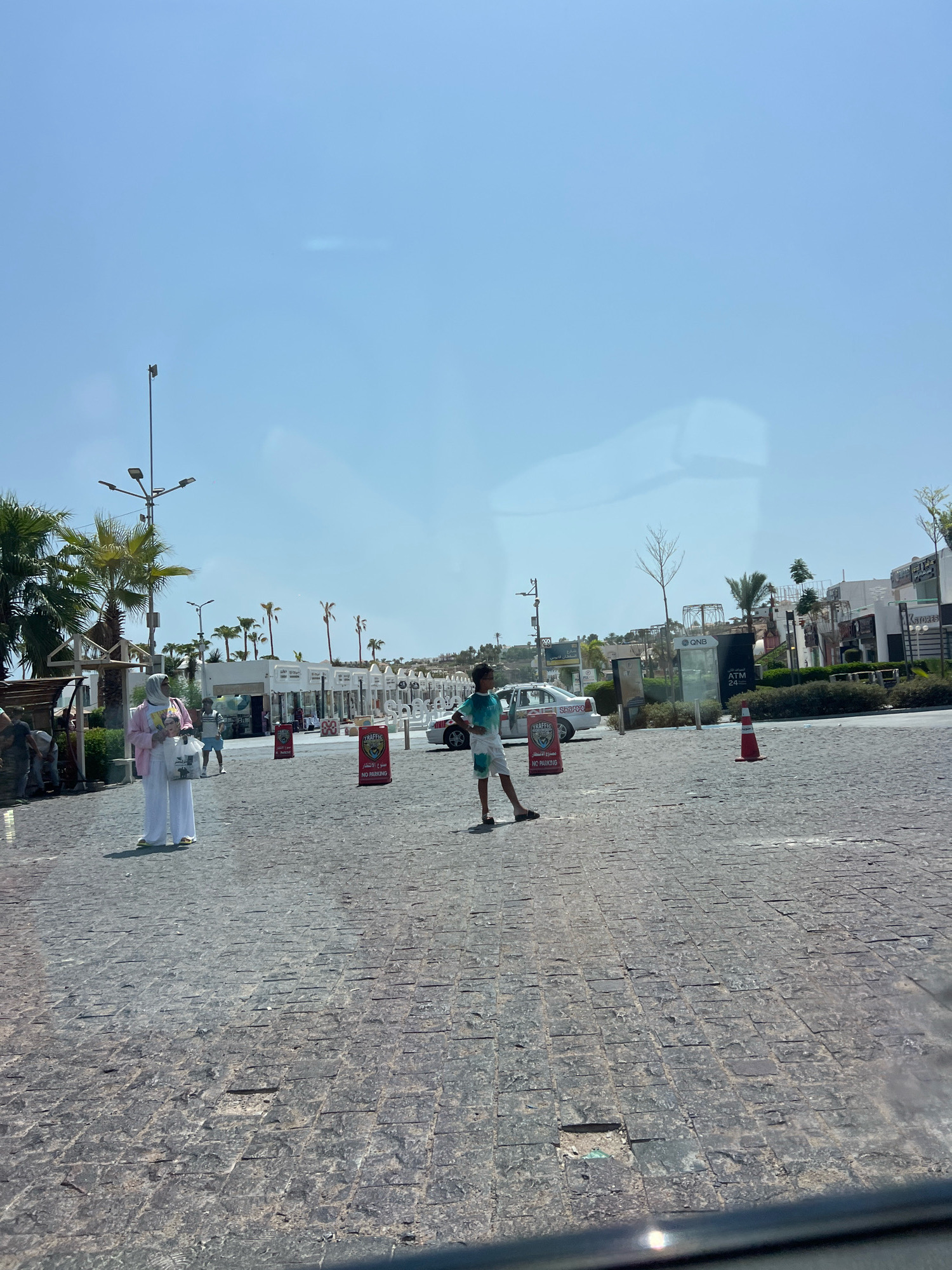 The image captures a street scene in Sharm El Sheikh, Egypt, on a clear, sunny day. The foreground is dominated by a textured, grey cobblestone pavement. In the middle ground, a young boy wearing a teal and white t-shirt and white shorts stands in the center of the pavement, looking towards the left. To his left, a woman dressed in a light pink abaya and a white hijab, carrying two shopping bags, walks towards the viewer. Behind them, a line of white buildings with arched entrances and signs, presumably shops and businesses, stretches into the distance. Palm trees are visible along the buildings and further back on a slight incline. A white car is parked near the buildings, and several "TRAFFIC NO PARKING" signs are placed on the pavement. The sky is a pale blue with a slight glare from the sun.