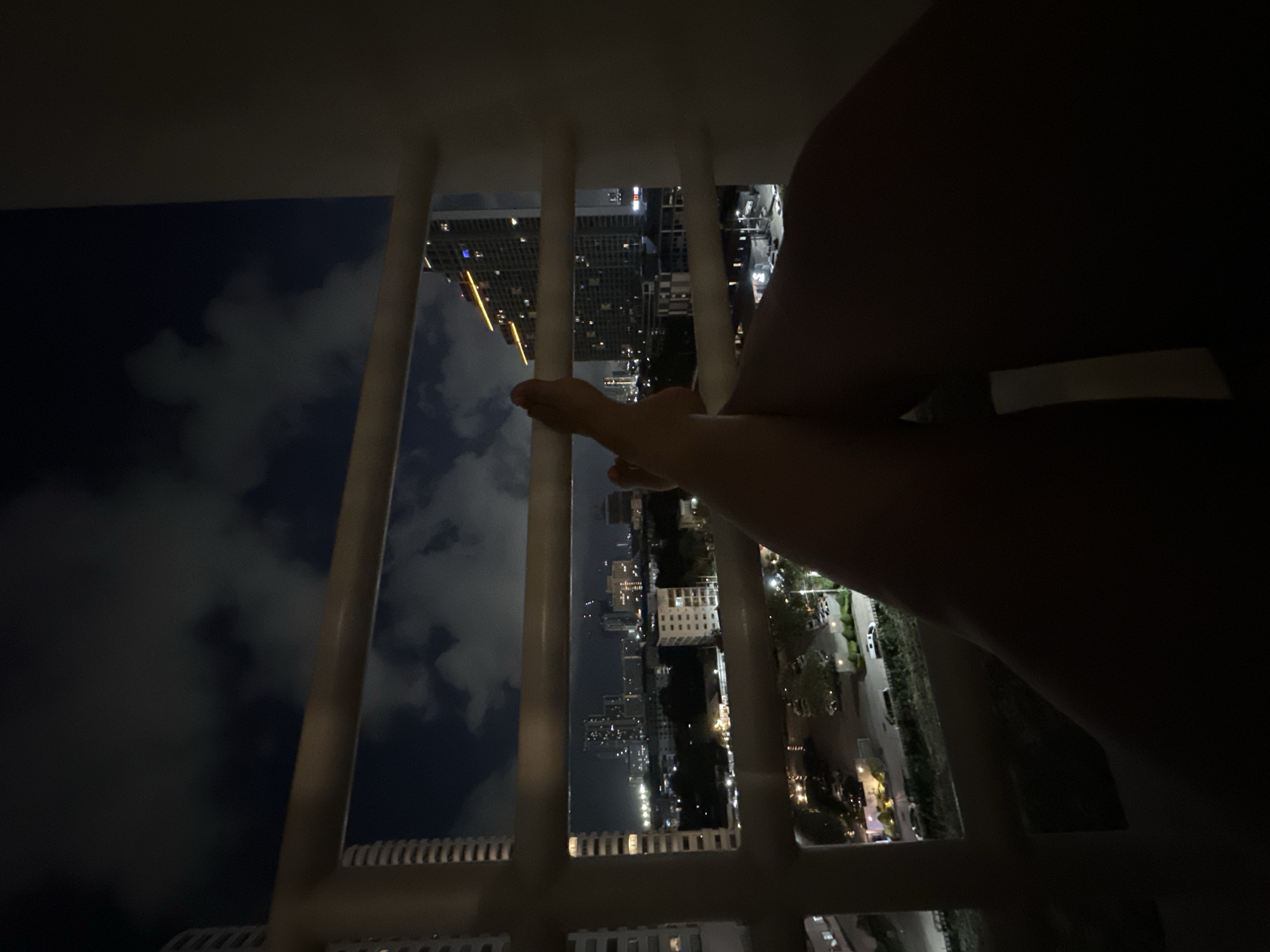 A night-time, indoor shot depicts a view of Pattaya City, Thailand, seen through white balcony railings. Tall buildings with illuminated windows are visible. Some clouds are present in the dark sky. A person's legs and feet are visible in the foreground, near the balcony railings. The perspective is angled downward.