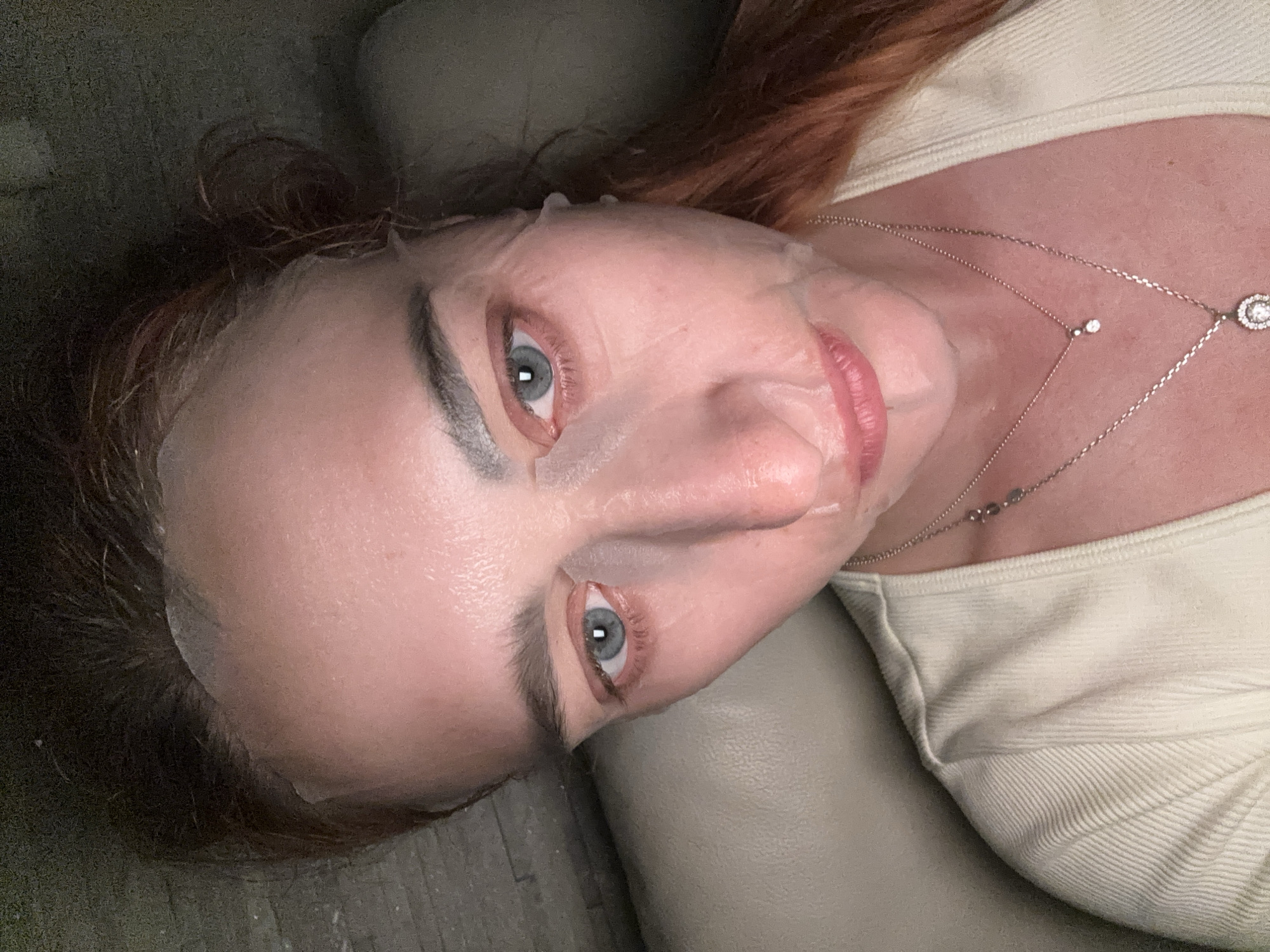 A woman is lying down wearing a facial mask. She has fair skin, gray-blue eyes, and reddish-brown hair. She is wearing a light-colored top and a delicate necklace. The setting appears to be indoors, possibly on a couch or padded surface. There is no indication of the location being Budva, Montenegro, or any specific city.