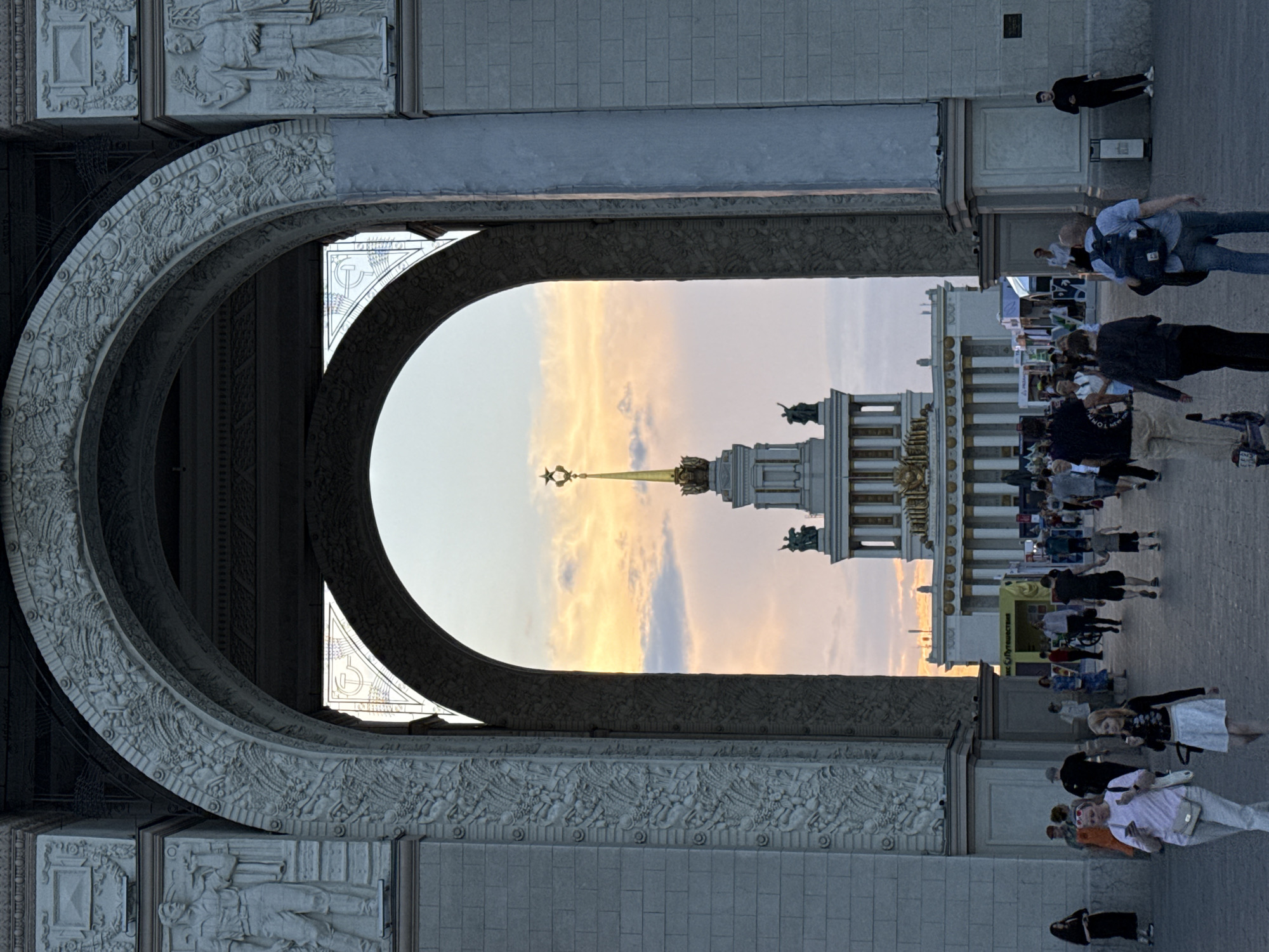 The image shows a view through an archway towards a large Soviet-era building topped with a golden star, located at VDNKh (Exhibition of Achievements of National Economy) in Moscow, Russia. The archway is elaborately decorated with bas-reliefs depicting agricultural and worker scenes. In the foreground, a crowd of people are walking on a paved area. The sky behind the building is filled with soft orange and pink clouds, suggesting it is either sunrise or sunset. The architecture is grand and monumental, characteristic of the Stalinist era.