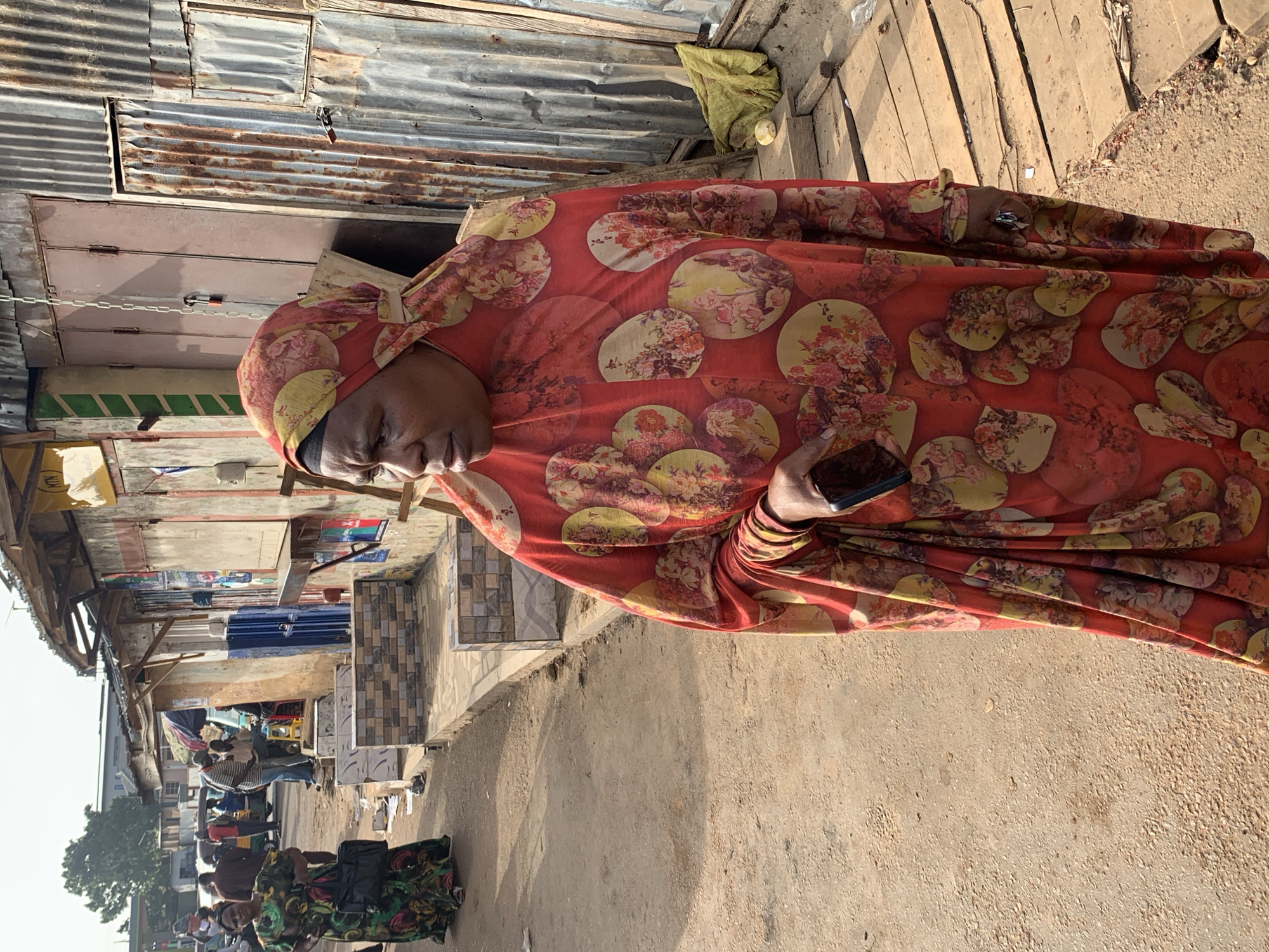 A woman wearing a red and floral patterned headscarf and a matching long dress stands in a street, looking to her left. She is holding a dark-colored mobile phone in her right hand. Behind her, a woman in a green and black patterned dress walks away from the camera, carrying a black bag. The background consists of a street with a dirt ground and several market stalls. One stall has a corrugated metal facade and wooden planks for a walkway. Other stalls are made of wood and have awnings. In the distance, there are people, vehicles, and more market structures. The lighting suggests it is daytime.