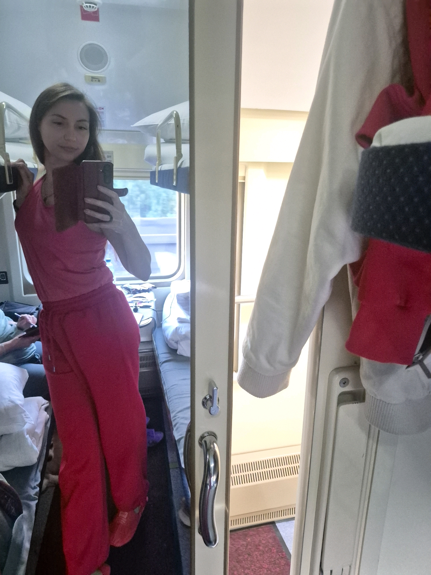 A young woman, dressed in a bright pink tracksuit and red slippers, stands in a narrow train compartment, taking a selfie in a mirror. She holds a dark red phone in her right hand, angled towards herself. The compartment features bunk beds with white bedding and metal railings, visible in the mirror's reflection. Outside the window, lush green trees can be seen passing by, indicating the train is in motion. To the right of the frame, a white hoodie and a dark blue and red garment are draped over a partition. The overall environment suggests travel within Russia, possibly on a long-distance train. The specific location in the city of Rodniki, Russia is not discernable from the visual information provided.